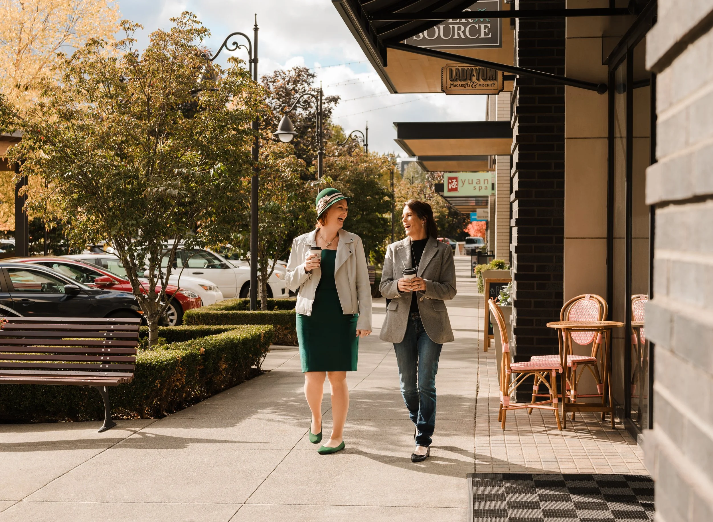 Two women walk and talk outside of a cafe on an autumn day, holding coffee cups, with trees and parked cars in the background.