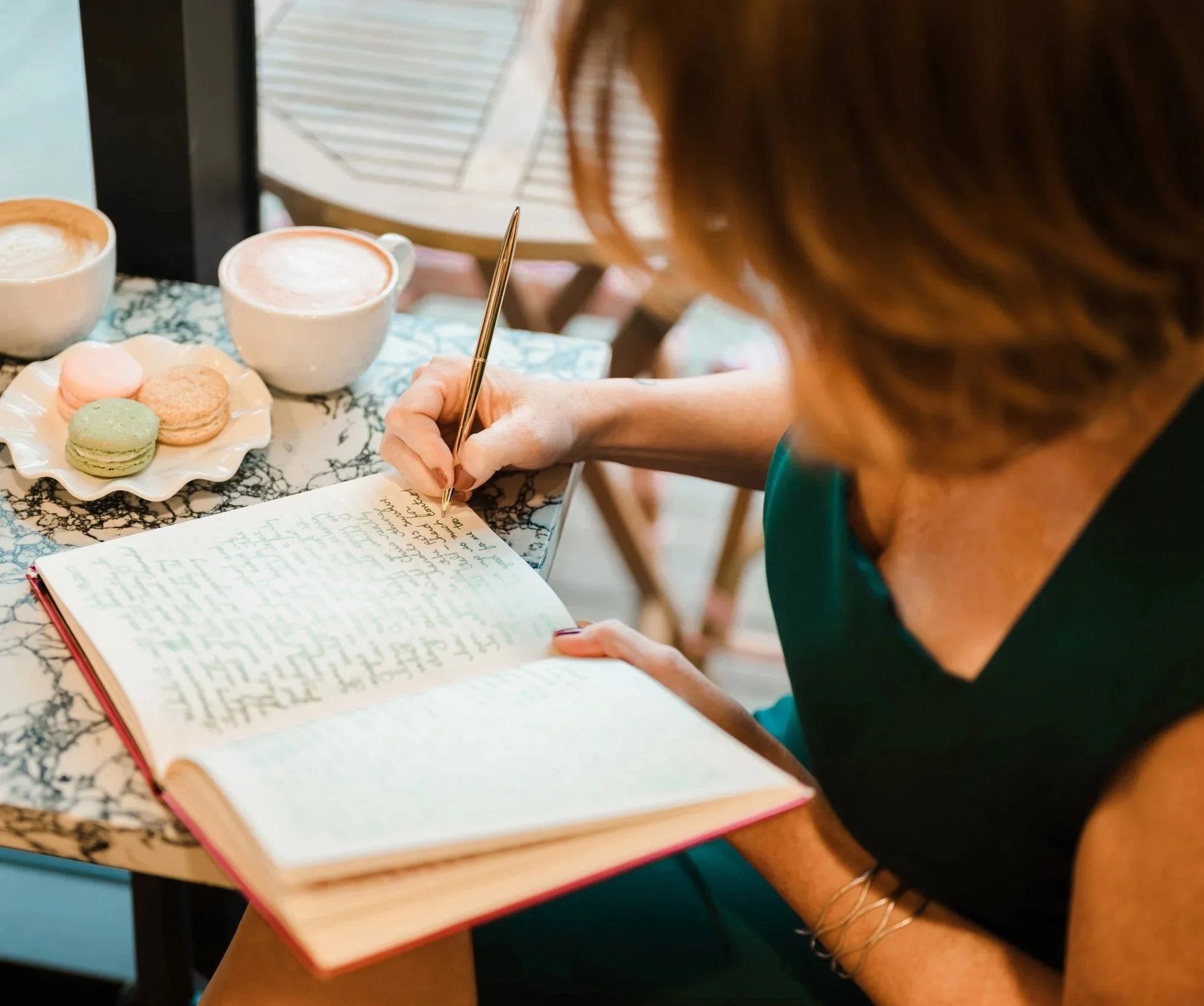A woman sitting at a table with two cups of coffee, a plate of macarons, and a notebook, writing with a gold pen.