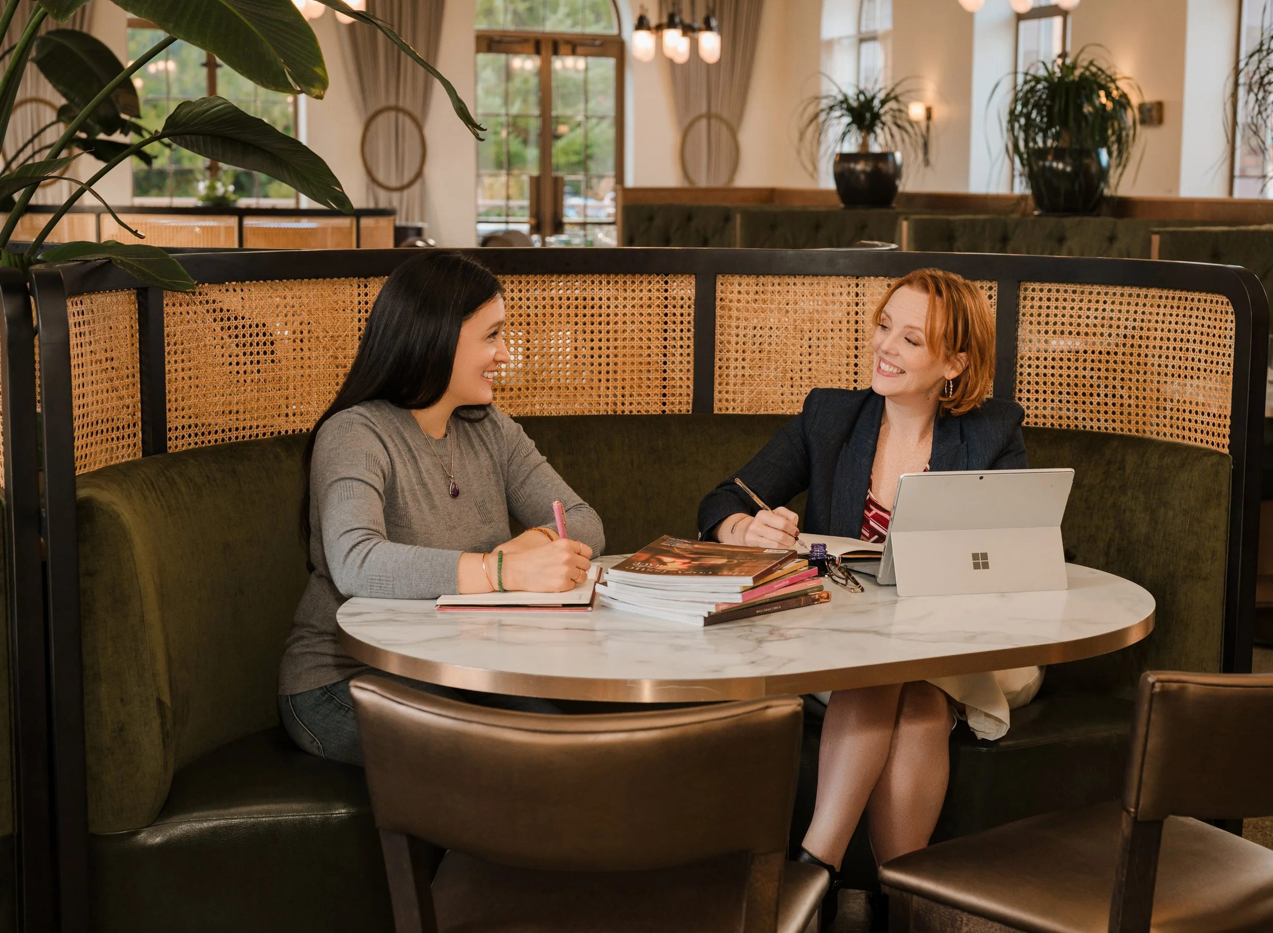 Two women sitting at a round marble table in a cozy restaurant or café, engaged in a lively conversation with notebooks and books in front of them, and a tablet on the table. They are smiling and appear to be having a friendly discussion.