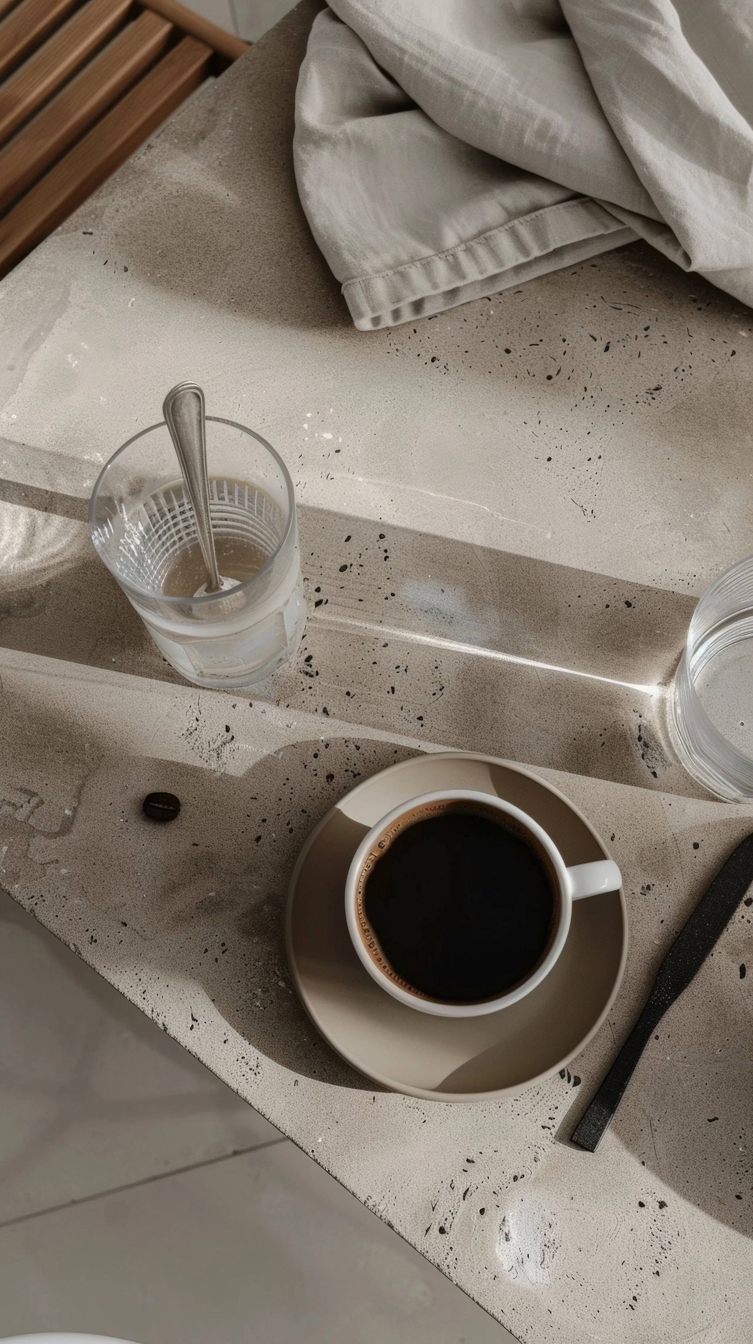 A beige countertop with a glass of water with a spoon, a cup of black coffee on a saucer, and a clear glass of water. There are some black specks on the surface and a white cloth and a black utensil nearby.