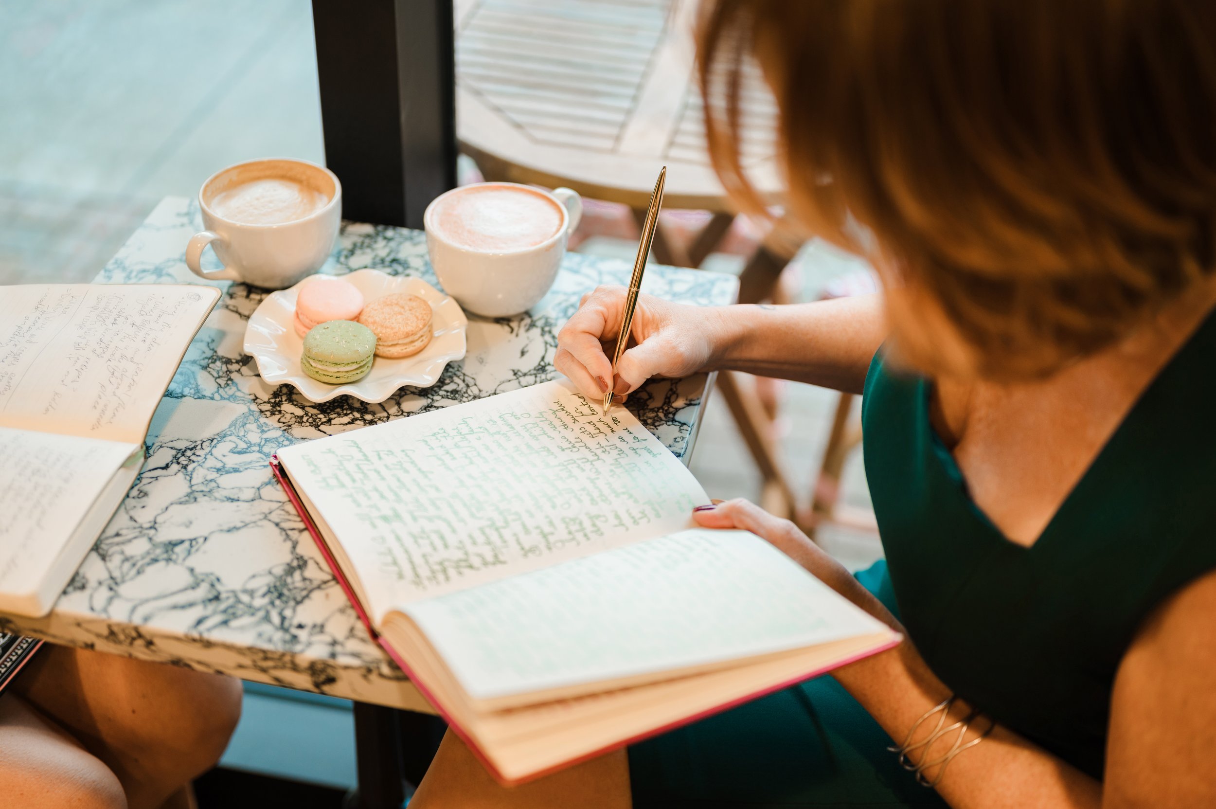 A woman sitting at a marble table, writing in a red notebook, with two cups of coffee and a plate of macarons on the table.
