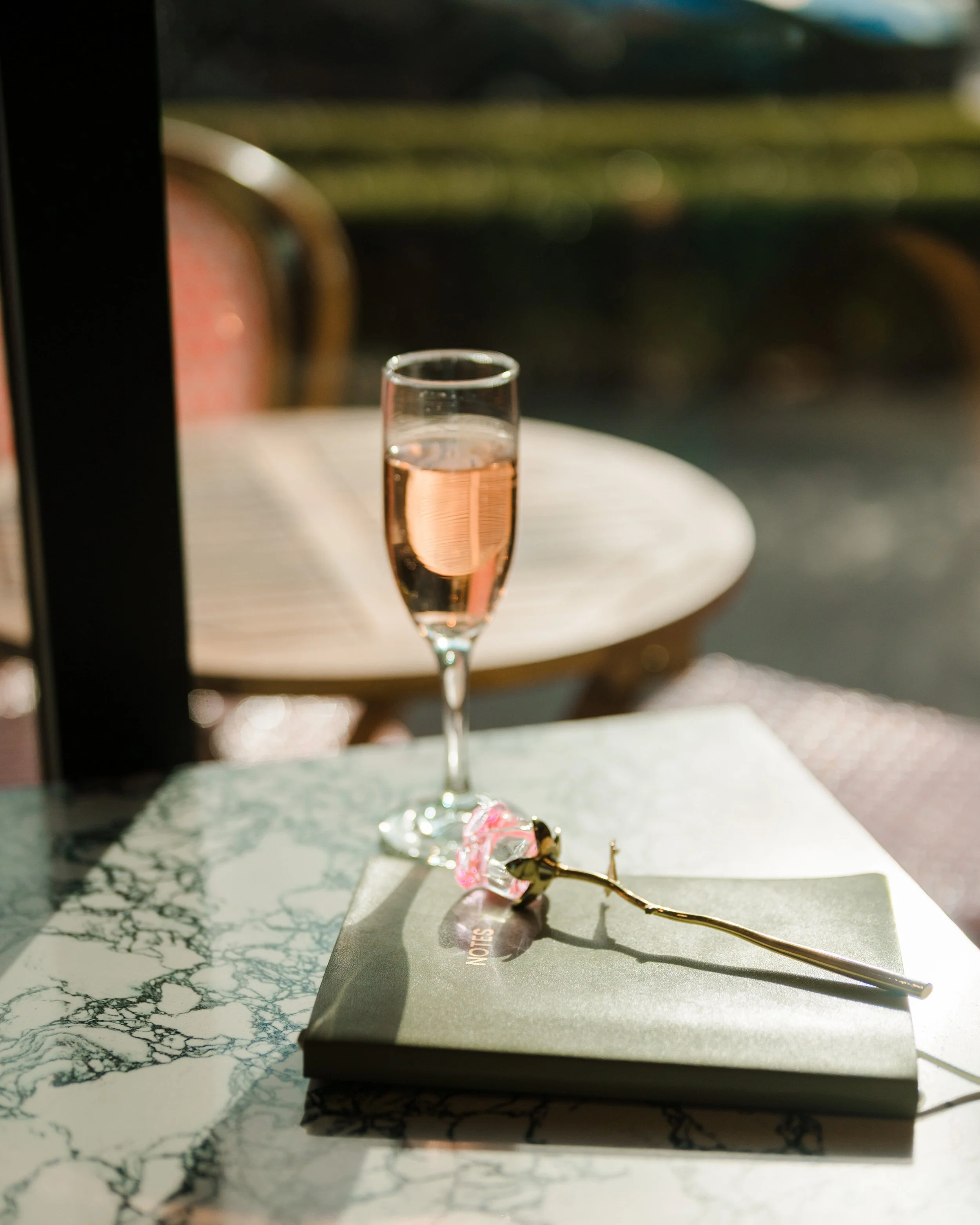 A glass of rosé wine on a marble table with a pink flower-shaped pen and a gray notebook.