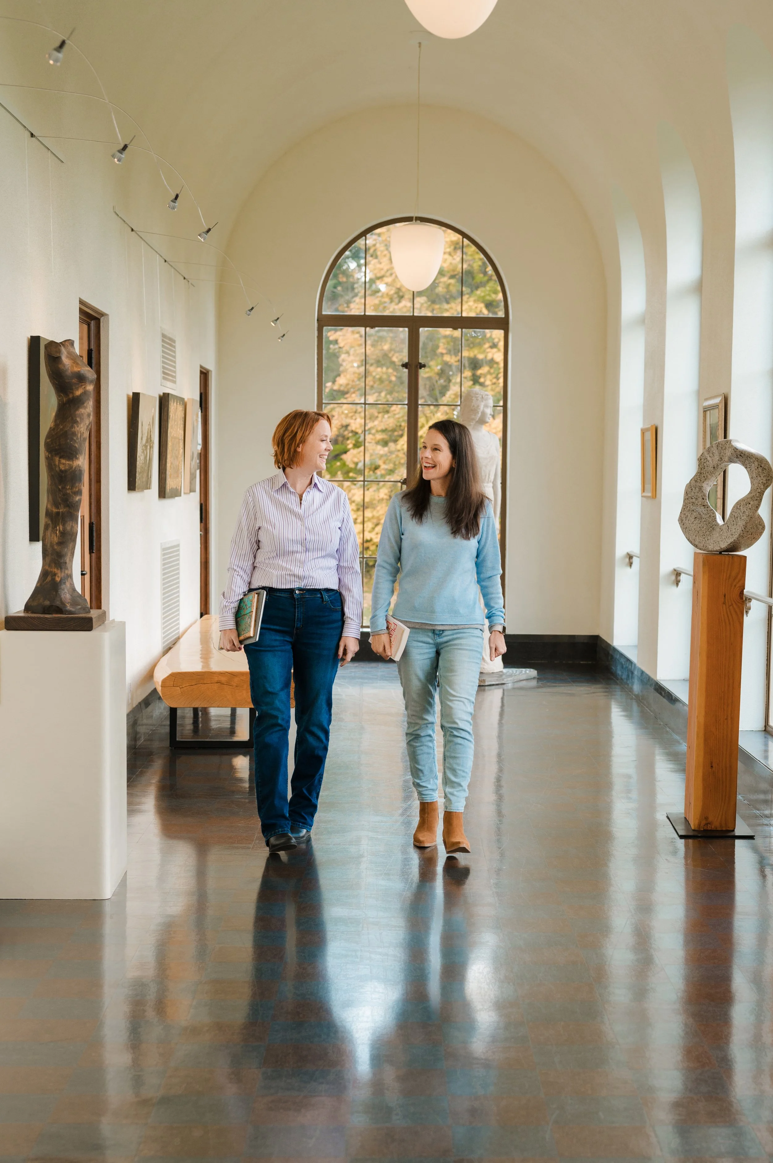 Two women walking and talking in an art gallery with sculptures and paintings, natural daylight streaming through large arched window.