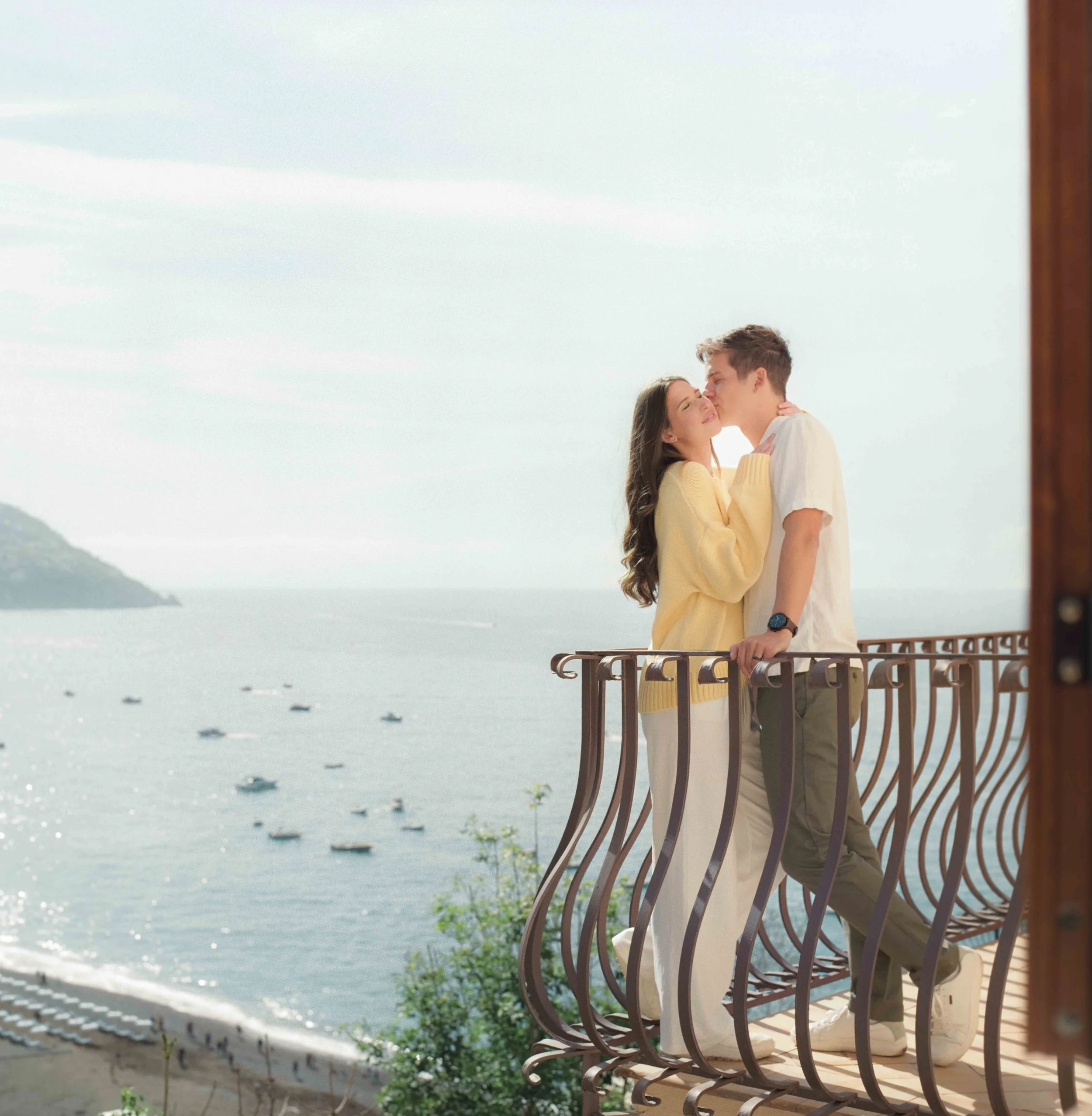 A couple sharing a kiss on a balcony overlooking the ocean with boats and distant land in the background Positano Italy