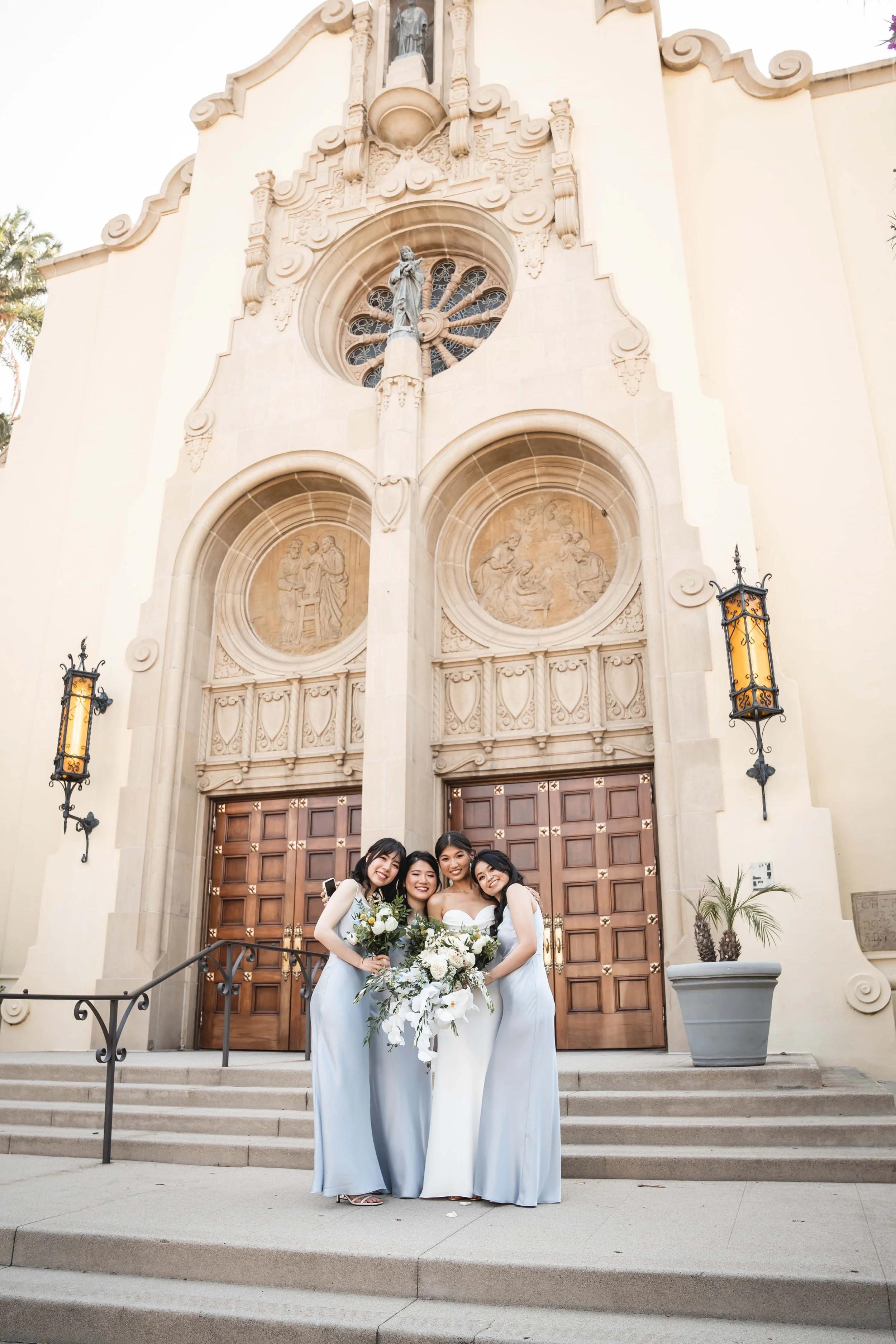 Group of four women in wedding dresses standing on church steps, holding bouquets, smiling.