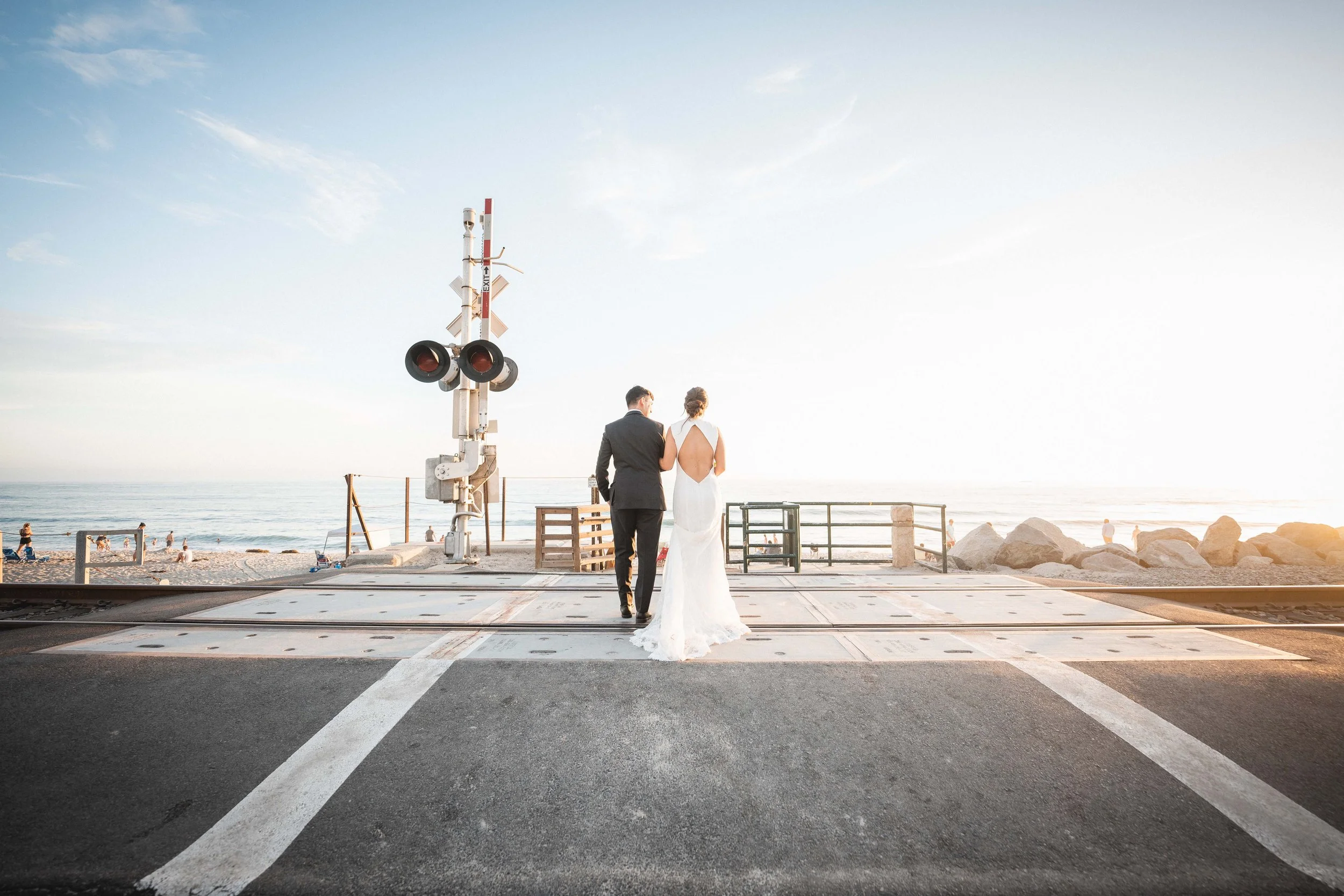 A bride and groom walking across a railroad crossing near the beach during sunset, with the sea and a few people in the background at Wedgewood San Clemente Shores