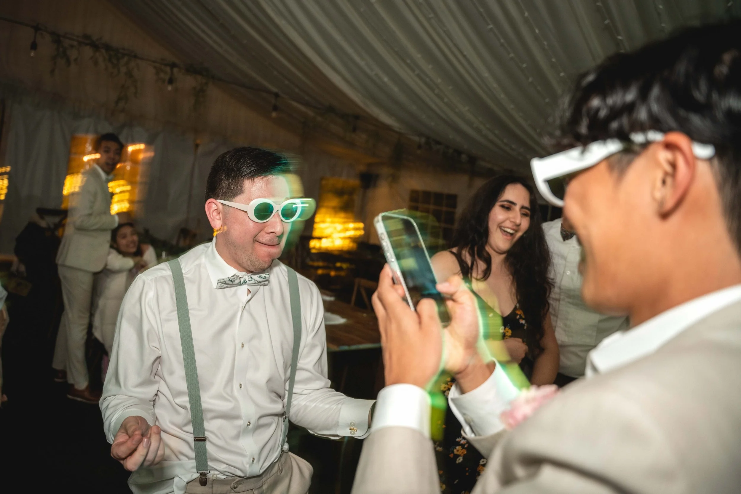 Group of people dancing and having fun at a party with one person wearing glowing sunglasses and a bowtie, another person holding a phone capturing the moment, and a woman laughing in the background inside a decorated venue, Botanica