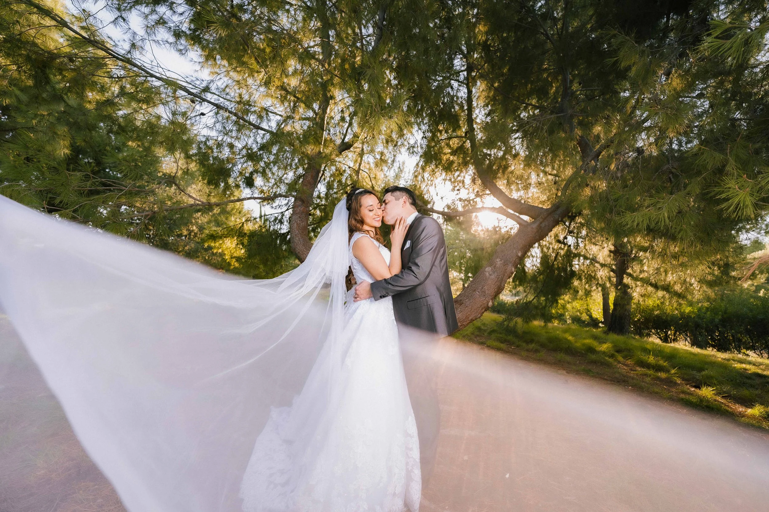 A bride and groom in wedding attire sharing a romantic kiss outdoors with trees in the background and sunlight filtering through the leaves, Jeffrey Open Space Trail