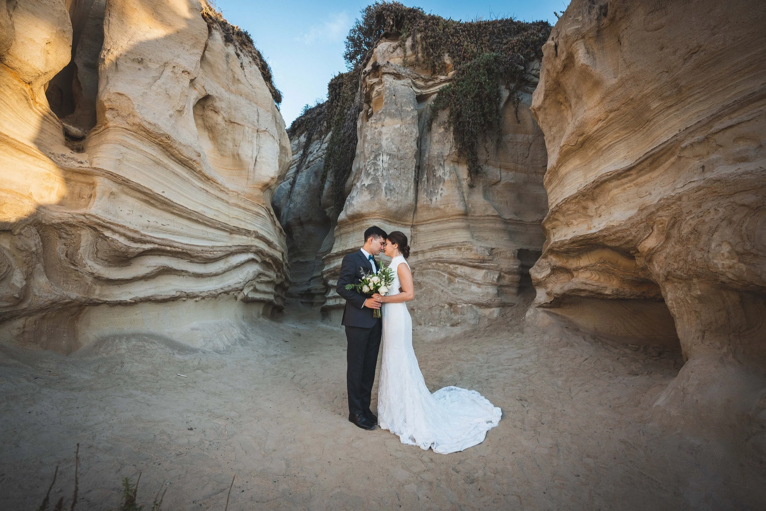 A bride and groom in wedding attire standing close together, holding hands and a bouquet of flowers, inside a natural rock formation or canyon with layered sandstone walls, San Clemente Shores Wedgewood