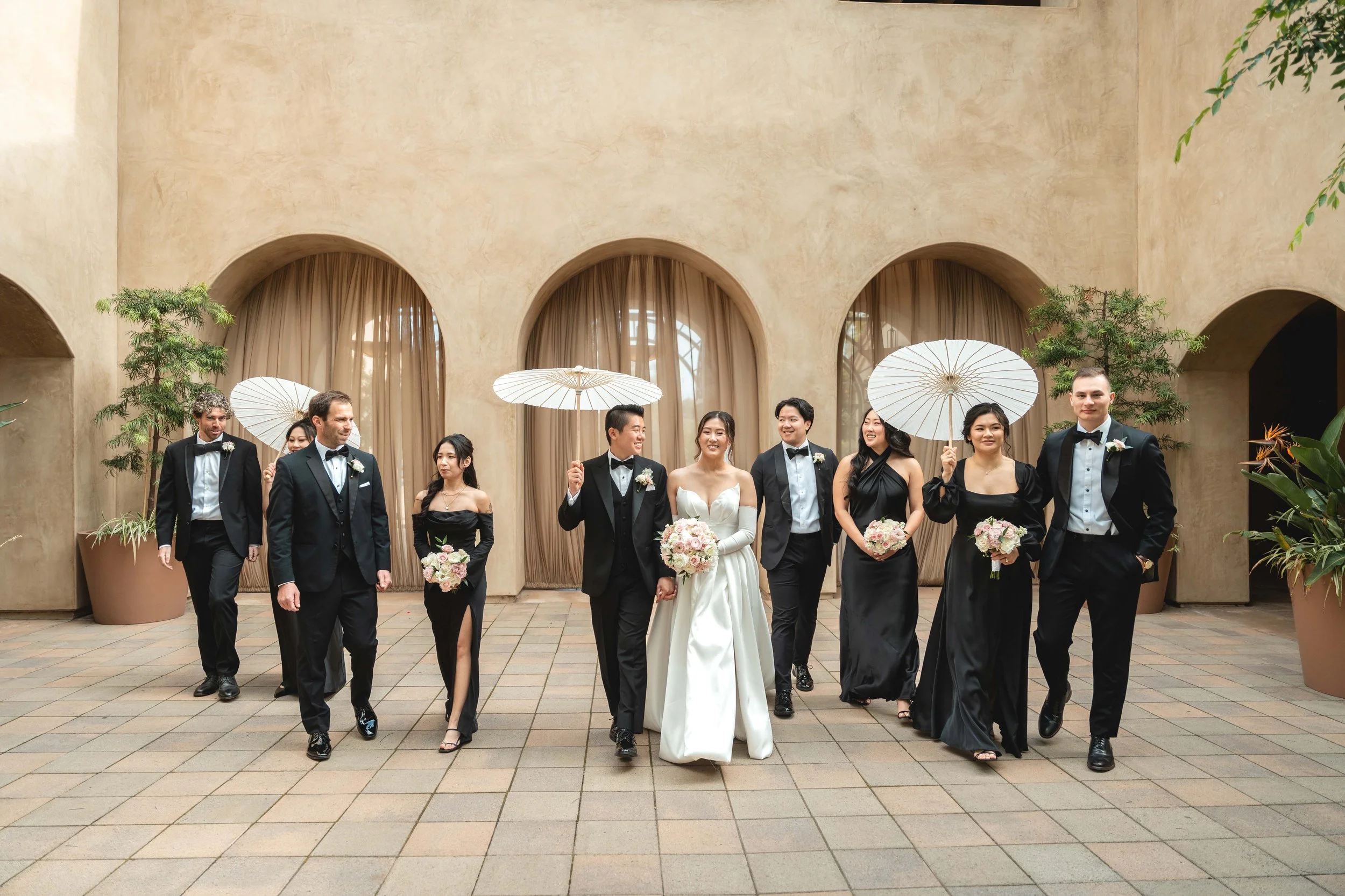 Wedding party with a bride in a white dress and groomsmen and bridesmaids dressed in black, walking outdoors with umbrellas and bouquets, in front of a beige building with arched windows and curtains, Serra Plaza