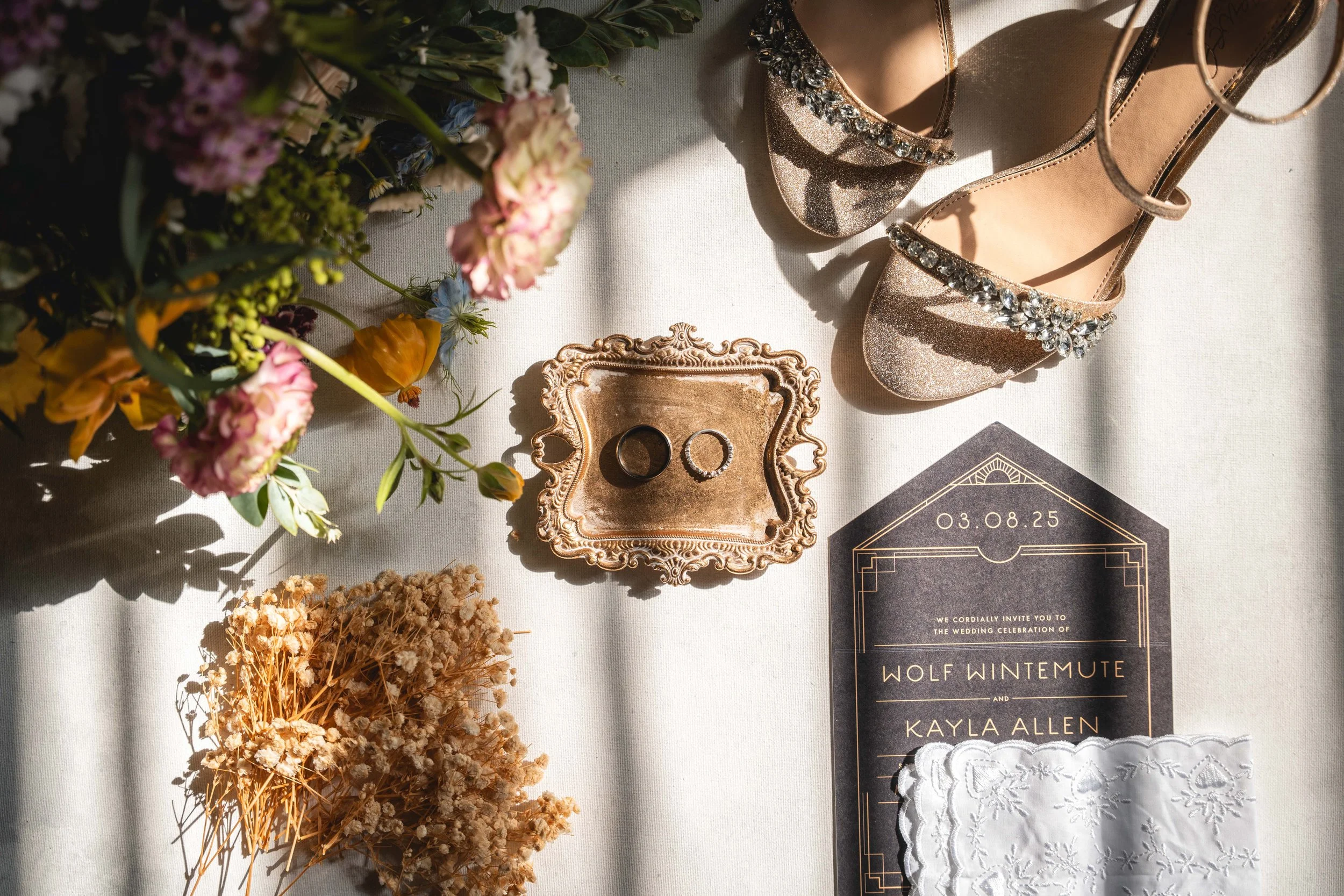 Flat lay of a wedding invitation, a pair of sparkly high-heeled shoes, wedding rings, dried flowers, fresh flowers, and a lace cloth on a white surface with sunlight casting shadows.