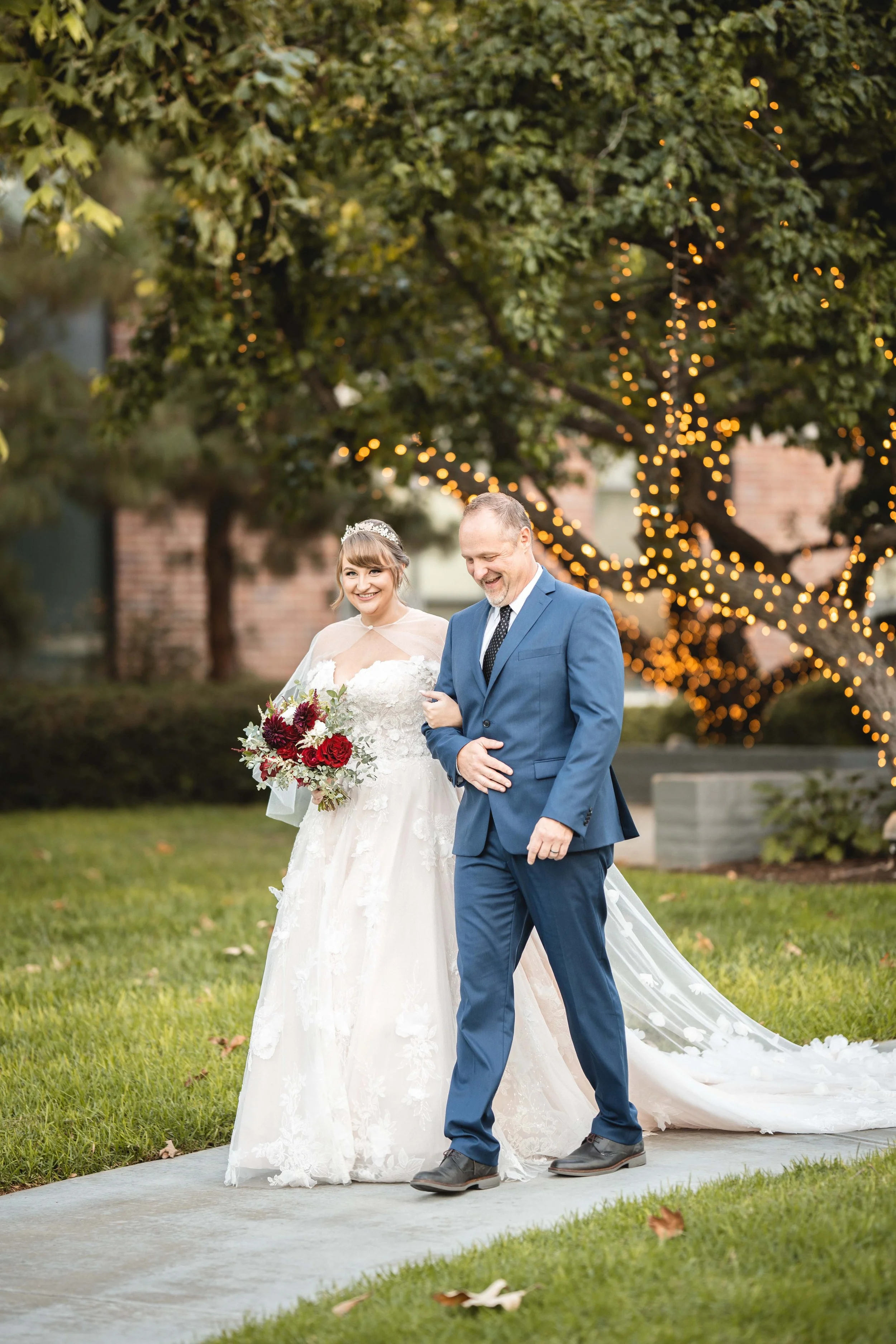 A bride in a white wedding gown and a tiara, holding a bouquet, walking arm-in-arm with a man in a blue suit, outdoors in a park with a lit, decorated tree in the background at Wedgewood University Club at UCI.