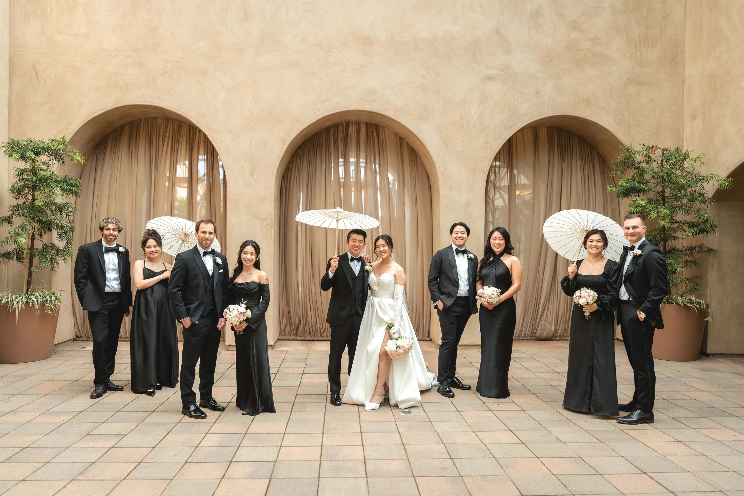 A wedding party with nine people, including bride and groom, standing in front of a beige wall with three arched windows and sheer curtains, some holding umbrellas, holding bouquets, all dressed in formal attire, Serra Plaza