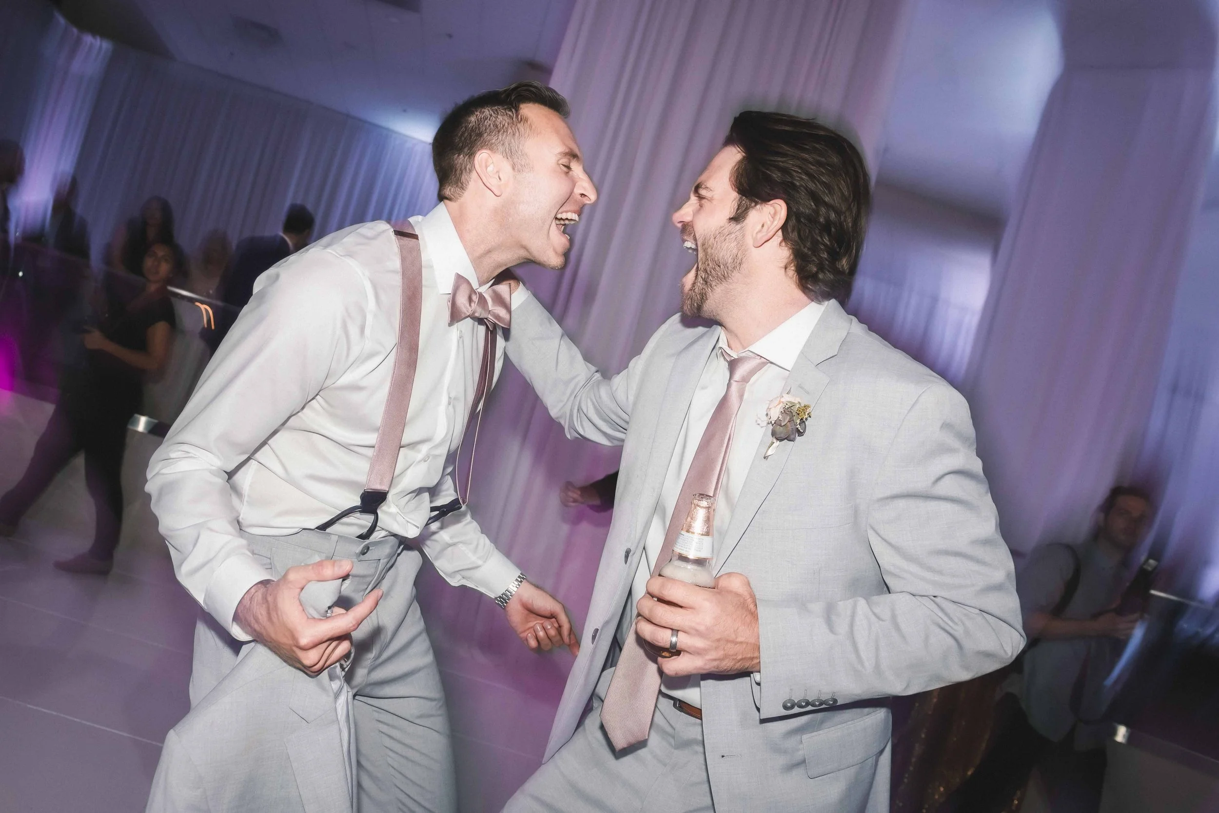 Two men in light gray suits, one with a pink bow tie and suspenders, the other with a pink tie and boutonniere, are joyful and laughing together at a wedding reception or party at JW Marriott in Palm Desert.