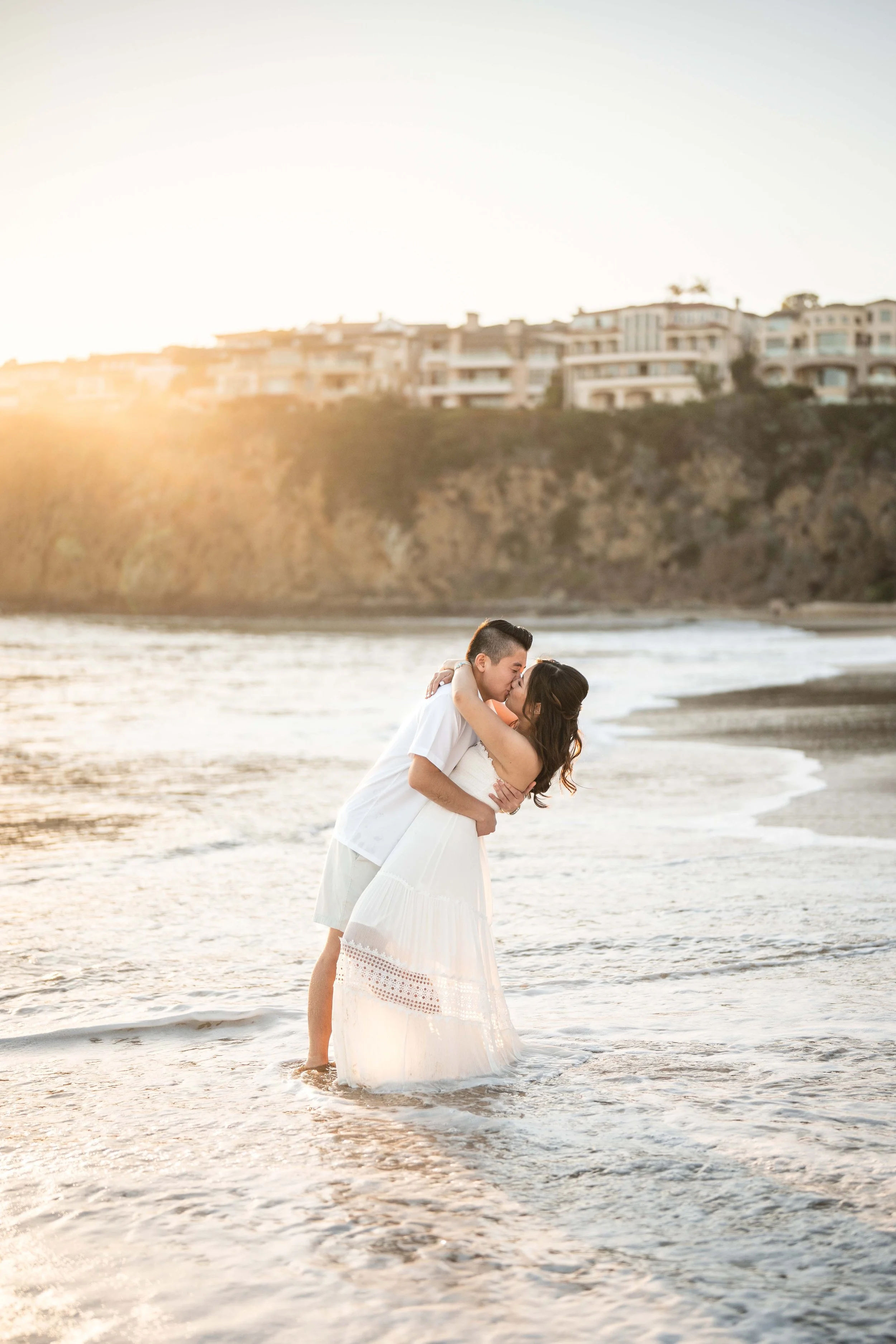 A couple in white attire sharing a kiss while standing in the shallow water at the beach during sunset, with buildings on a hill in the background, Laguna Beach