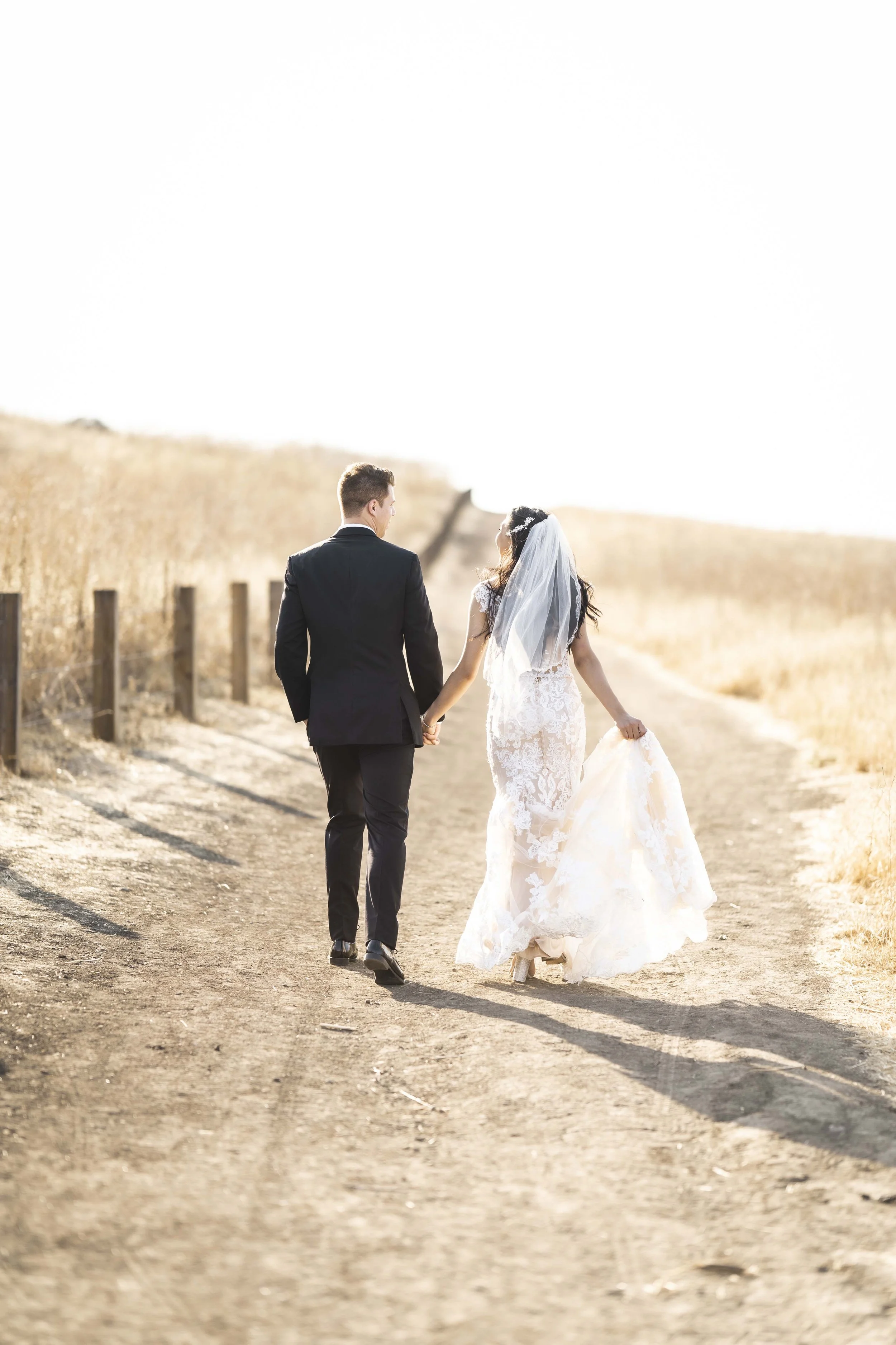 A bride and groom walking hand in hand down a dirt path in a rural area, with tall golden grass on either side, during sunset, Quail Hill