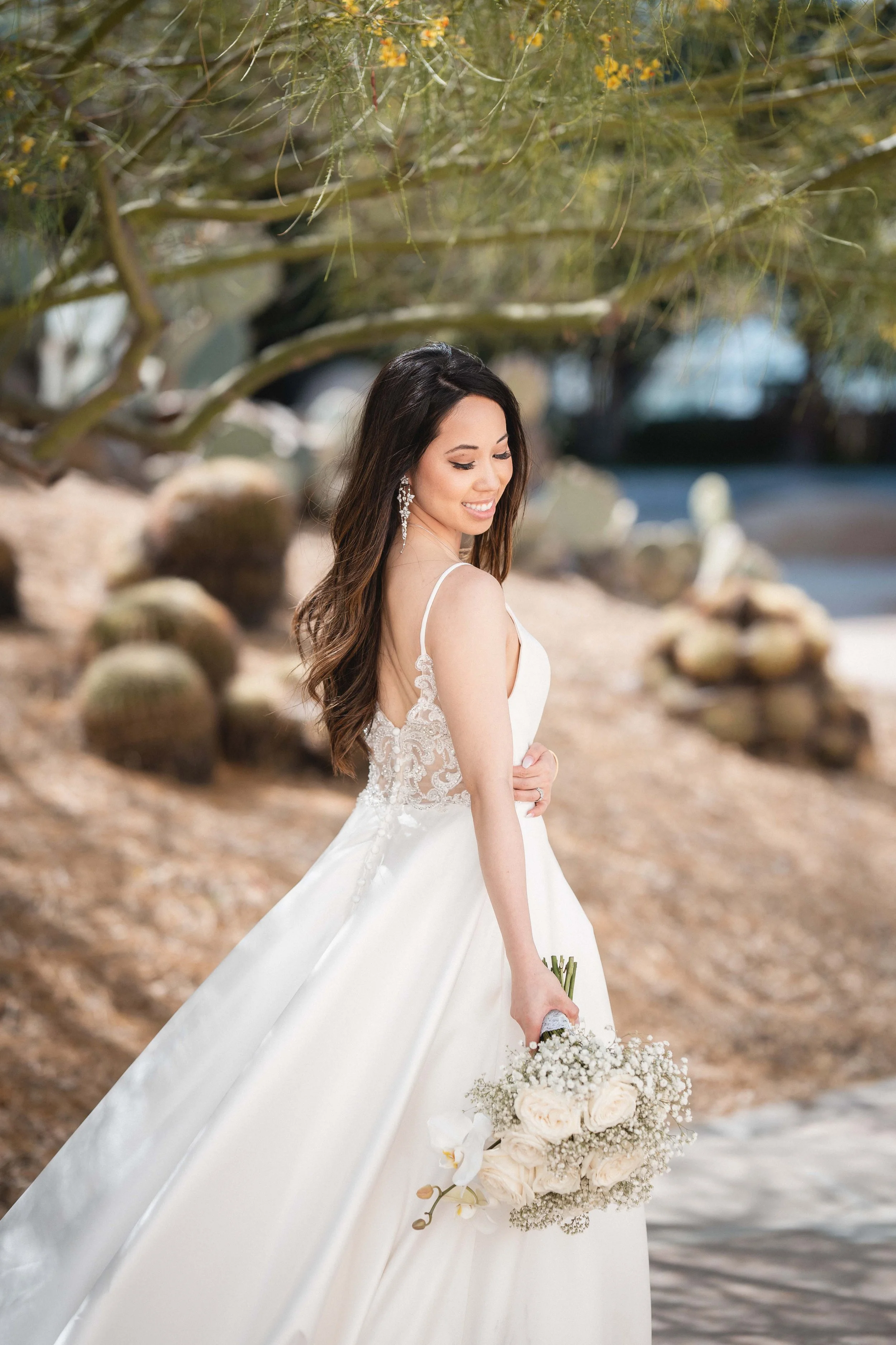 A woman in a white wedding dress holding a bouquet of white and cream flowers, standing outdoors near water with trees and rocks in the background, Noguchi Gardens