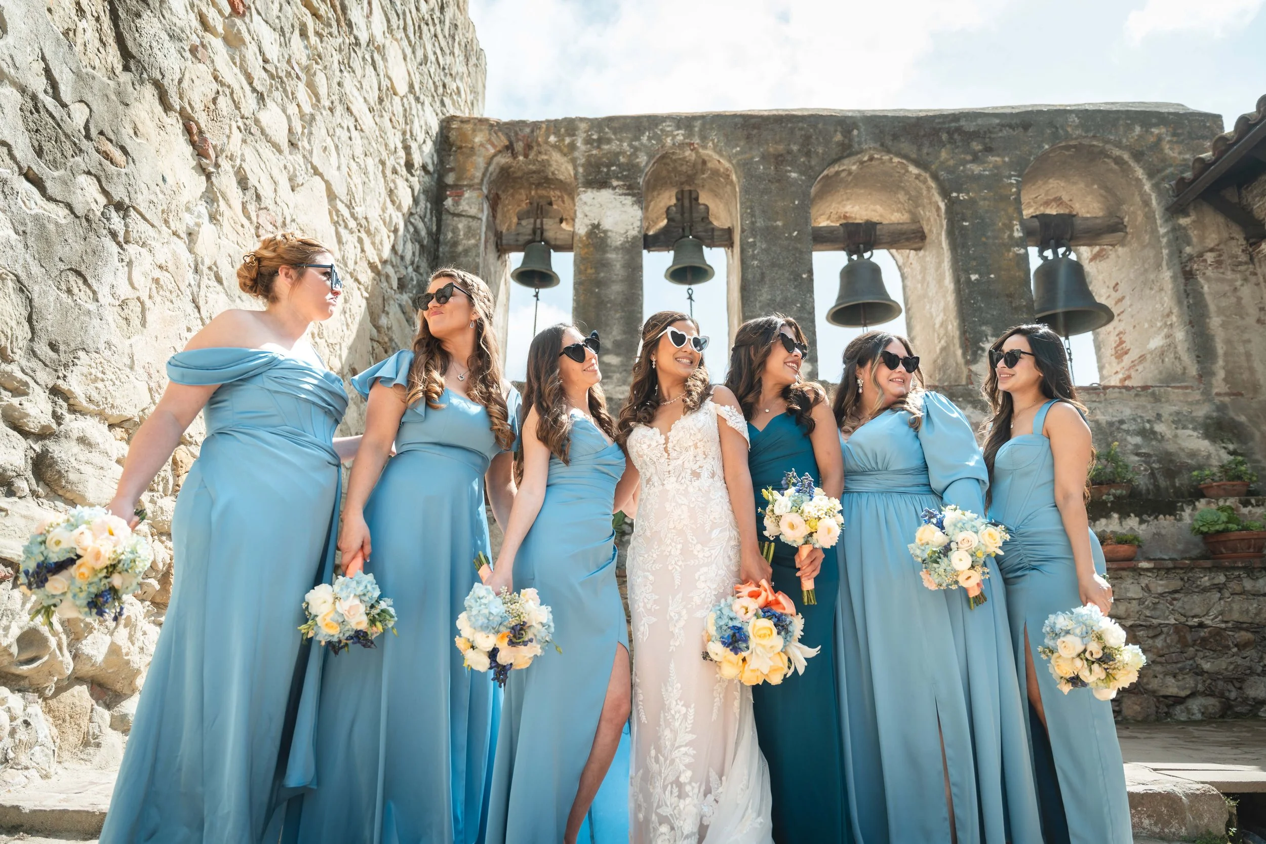 Bridal party of eight women in blue bridesmaid dresses and one bride in white wedding dress, holding bouquets, standing outdoors near an ancient stone wall and bells, on a sunny day, Mission San Juan Capistrano