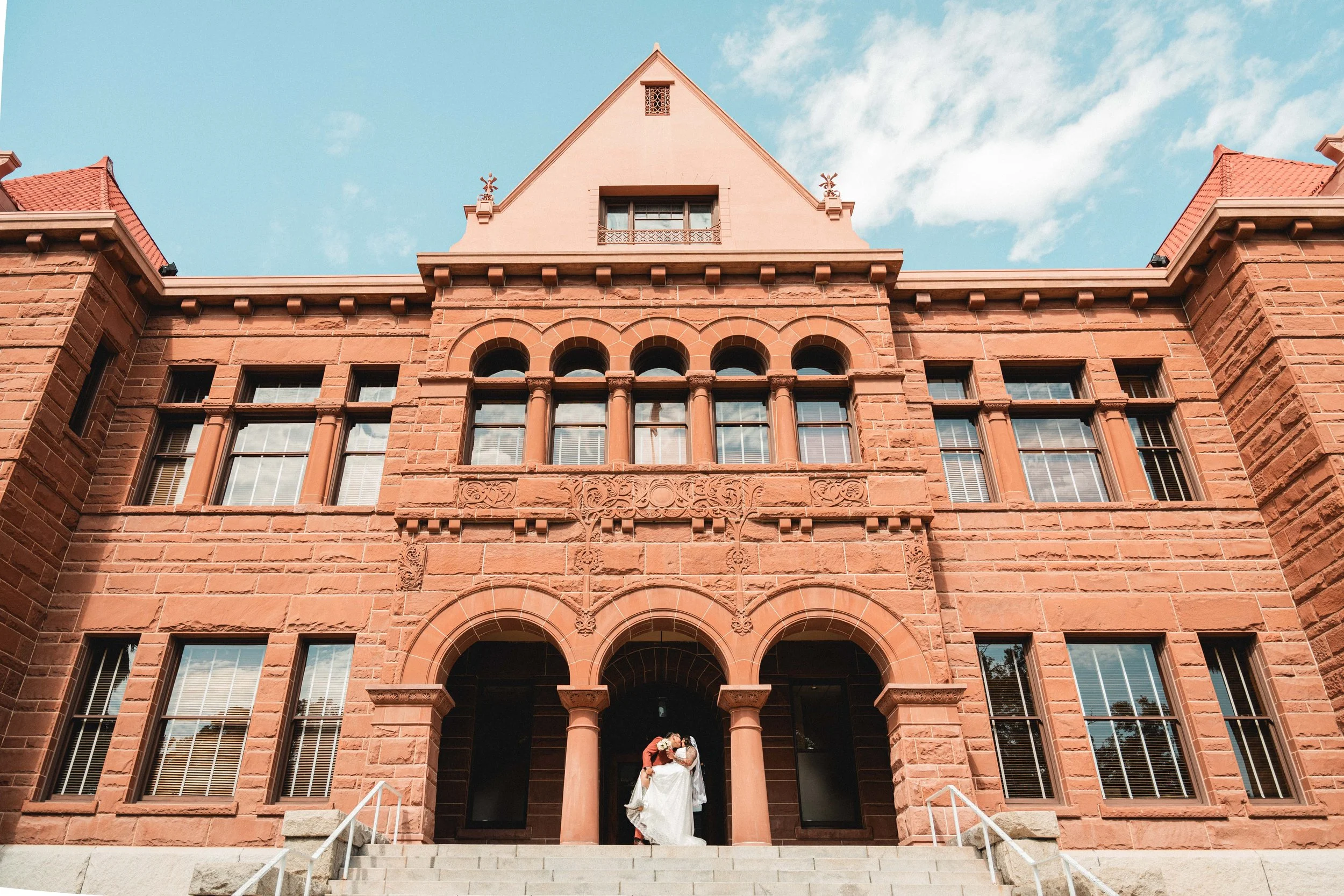 A bride and groom standing on the steps of a historic red brick building with arched windows and columns, under a partly cloudy sky, Orange County Courthouse