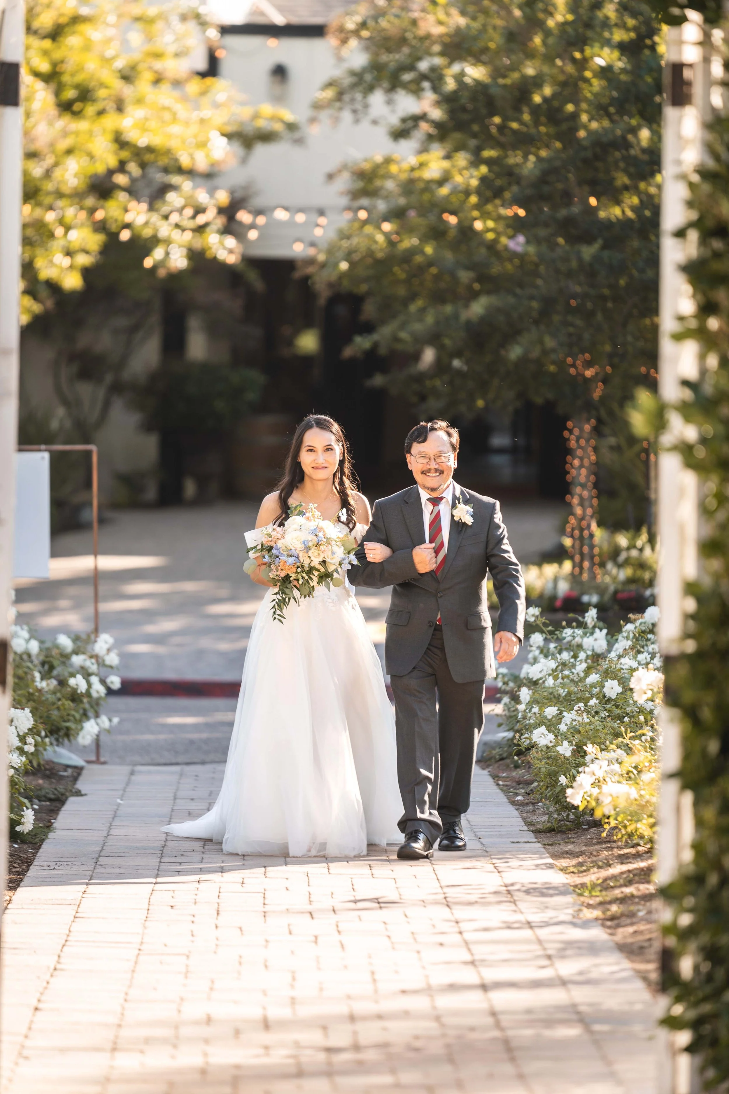 A bride in a white wedding dress holding a bouquet walking down an outdoor aisle with an older man in a dark suit and striped tie, smiling, during a wedding ceremony in a garden setting with trees and white flowers, Coto Valley Country Club