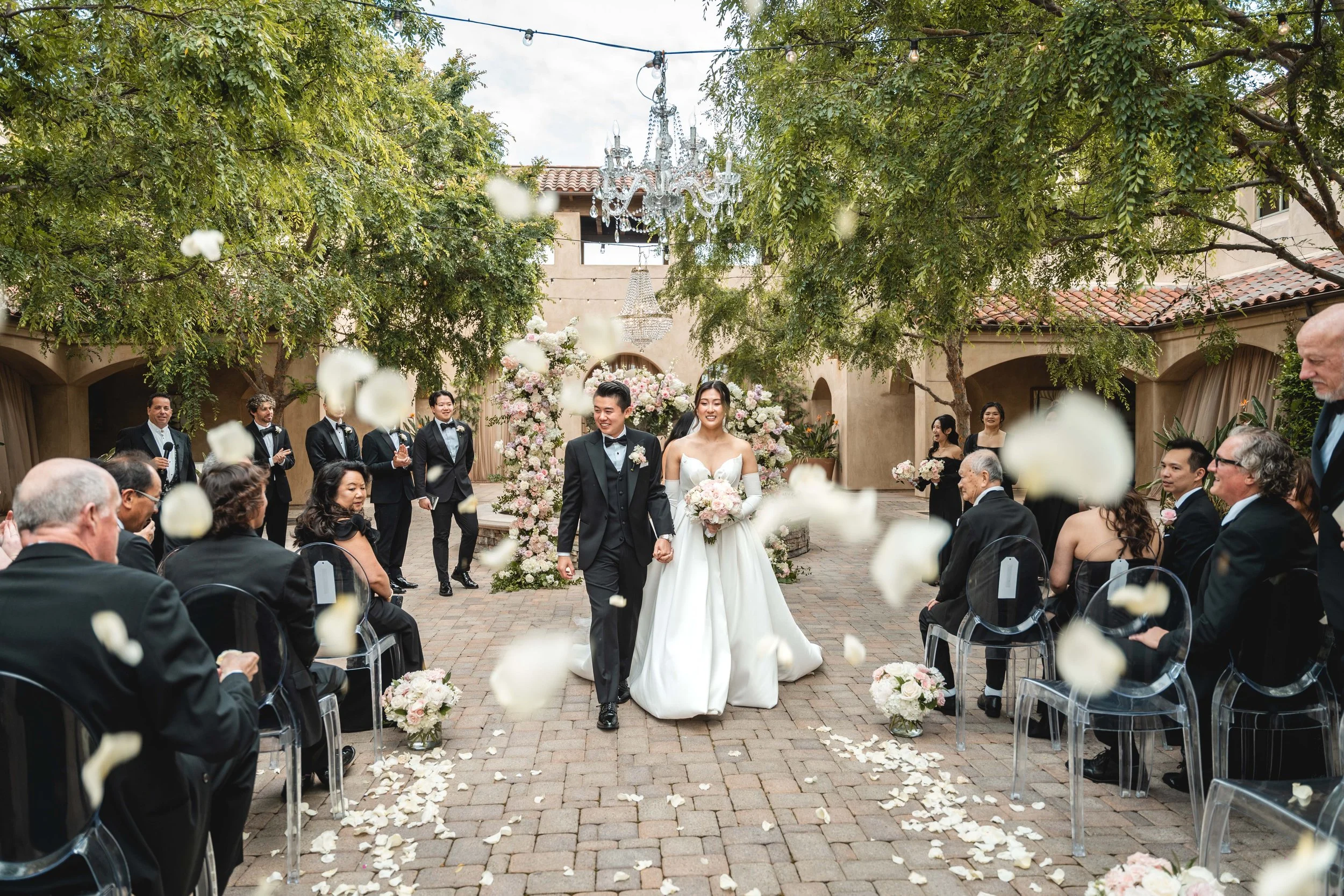 A bride and groom walk down the aisle at an outdoor wedding ceremony, surrounded by guests seated on either side and wedding party members standing behind them, with flower arrangements and petals on the ground, under a canopy of trees, Serra Plaza