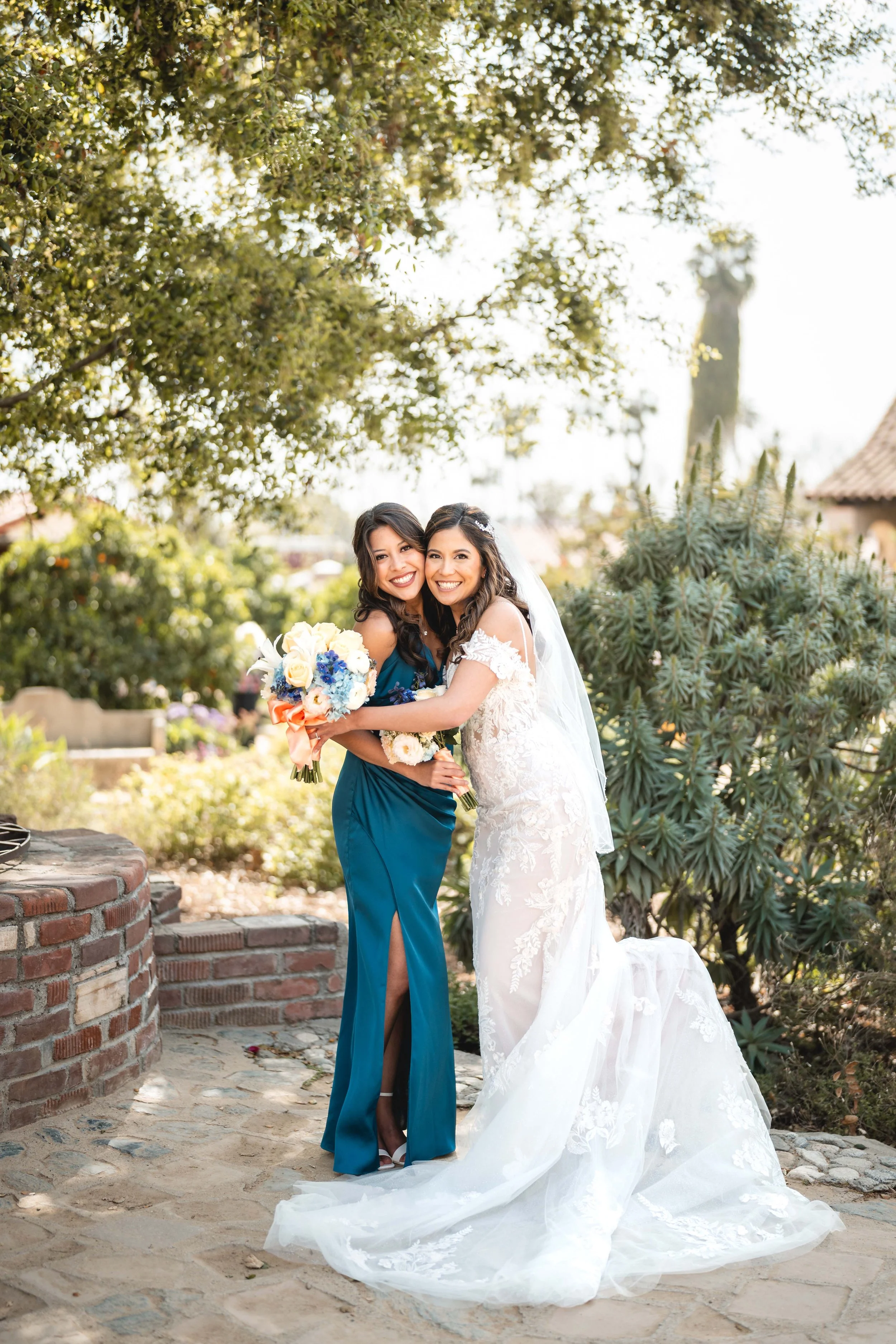 A bride in a white wedding gown and a woman in a teal dress hugging, holding a bouquet, outdoors with trees and plants, Mission San Juan Capistrano