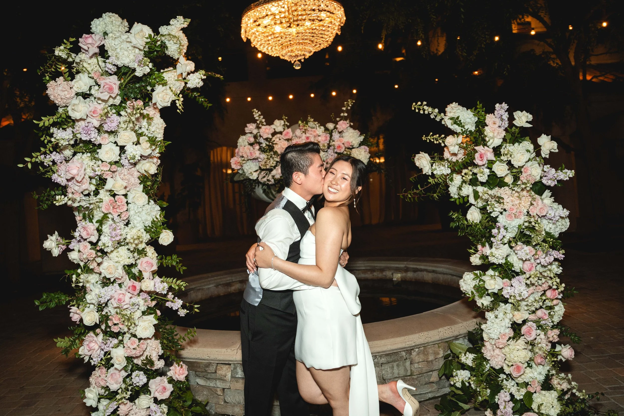A happy couple on their wedding night, posing under a floral arch with pink and white flowers at night, with a chandelier and string lights in the background, Serra Plaza