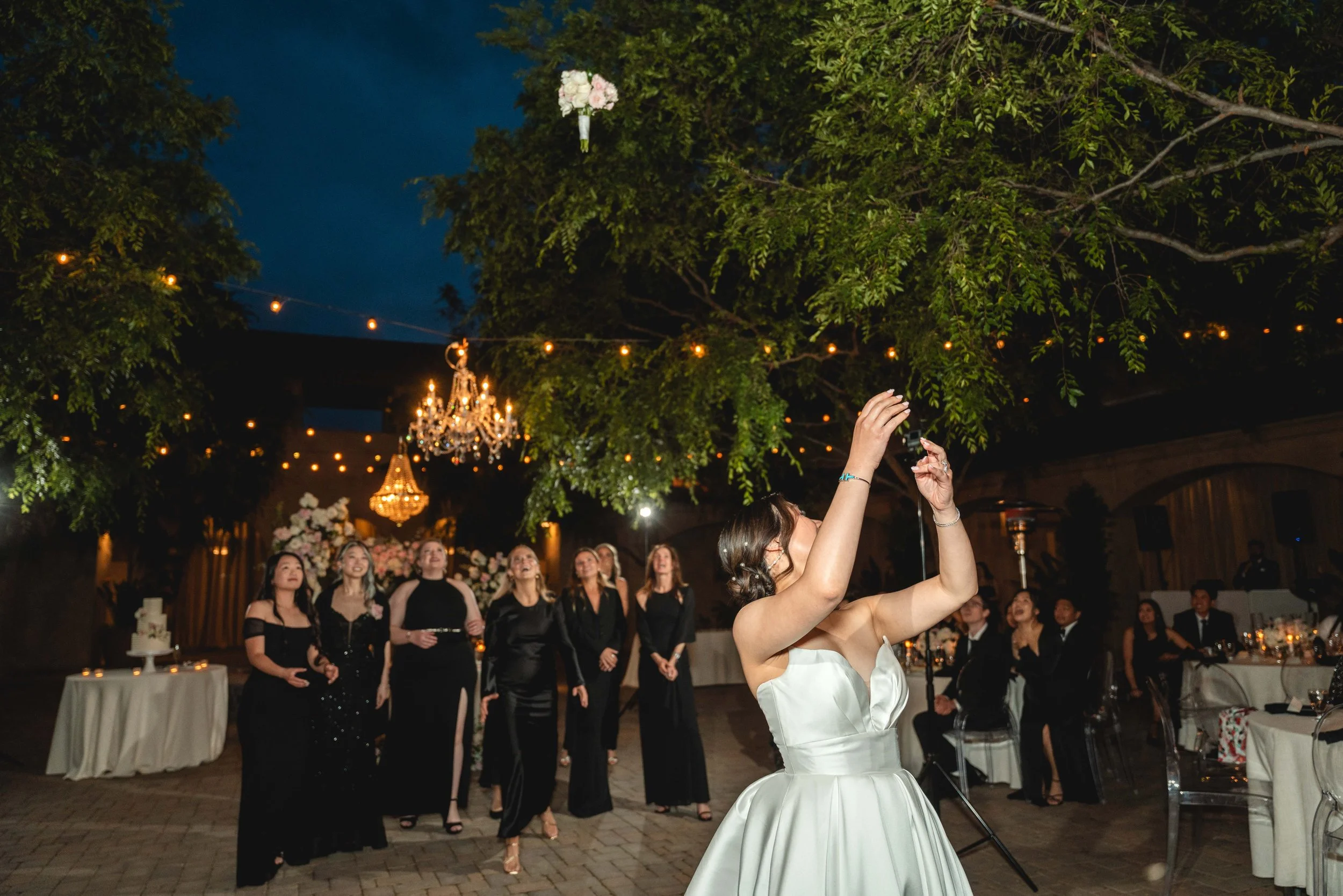 A woman in a white wedding dress is throwing her bouquet to a group of women at her wedding reception, which is decorated with chandeliers, lights, and floral arrangements outdoors at night, Serra Plaza