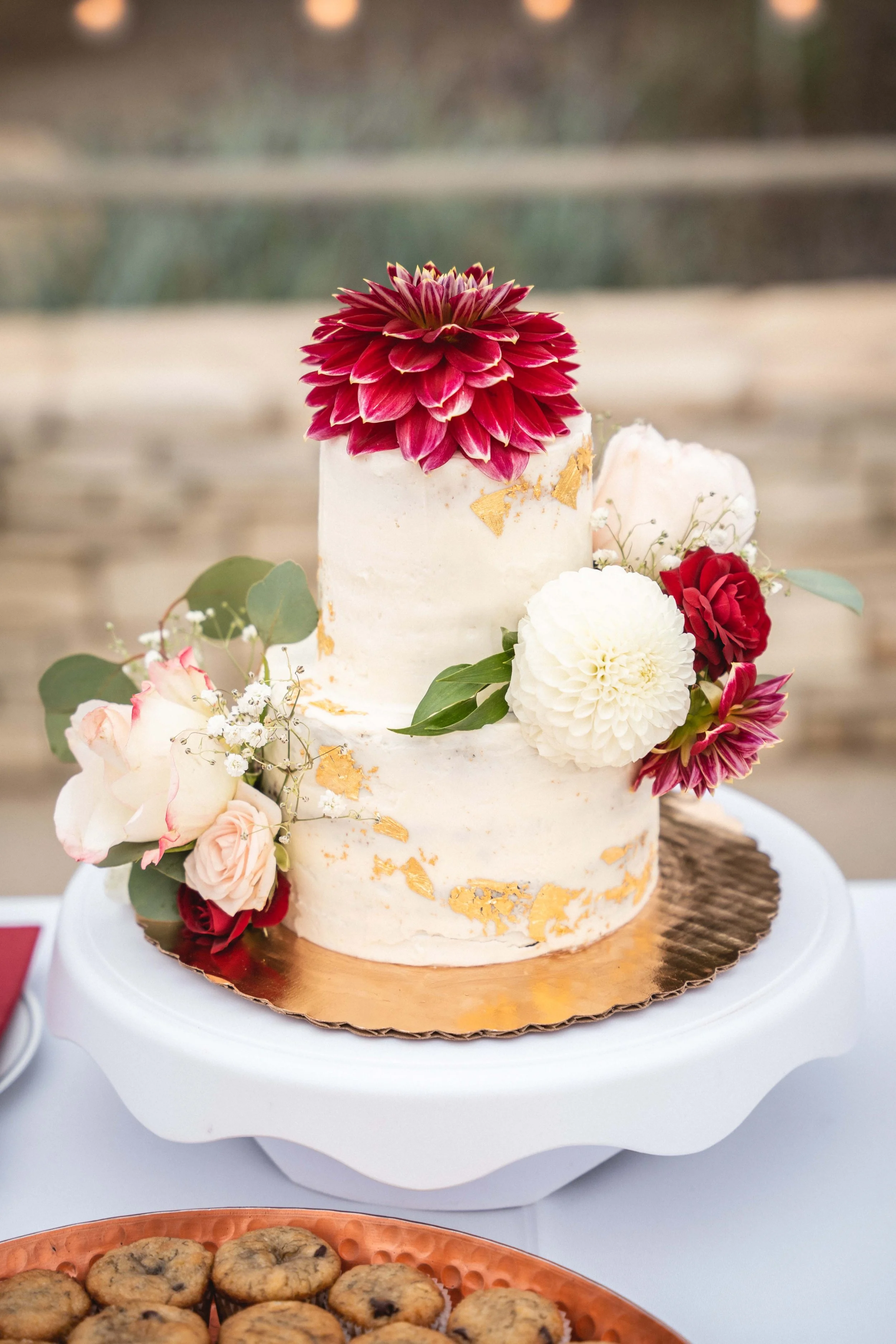 A cake decorated with pink and white flowers and gold leaf, displayed on a white cake stand at a celebration event at Point Vincent Interpretive Center