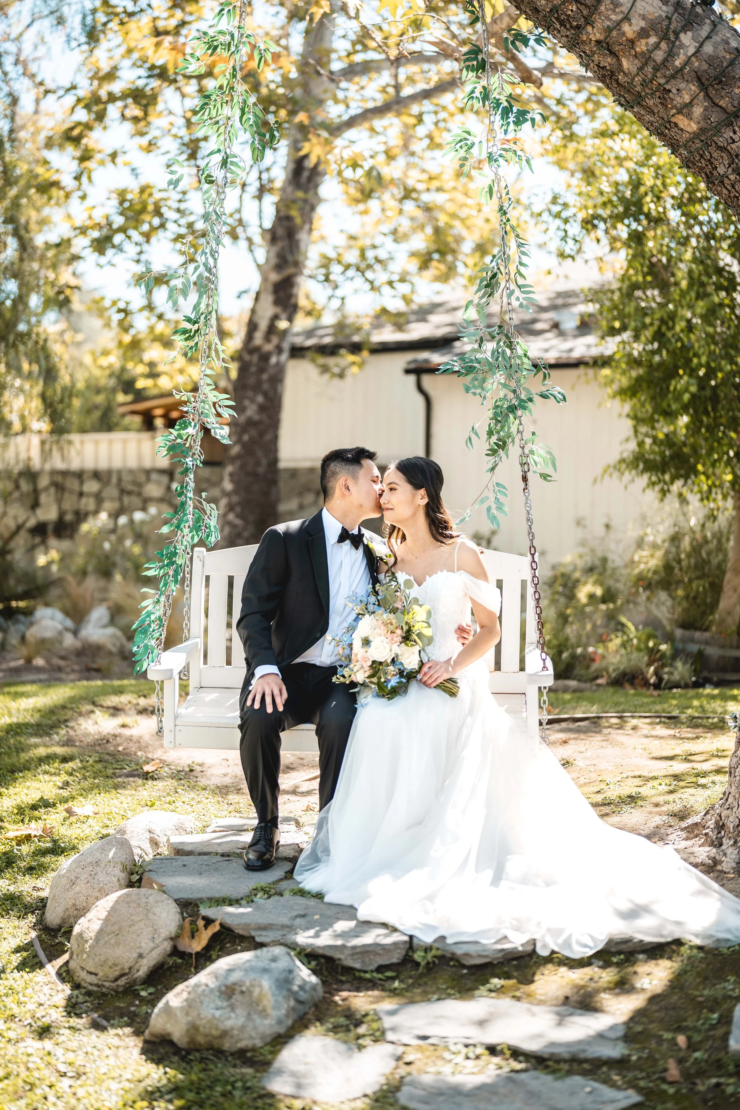 A newlywed couple sitting on a white wooden swing in a garden, with the bride holding a bouquet and the groom in a black tuxedo, sharing a kiss in the sunlight, Coto Valley Country Club