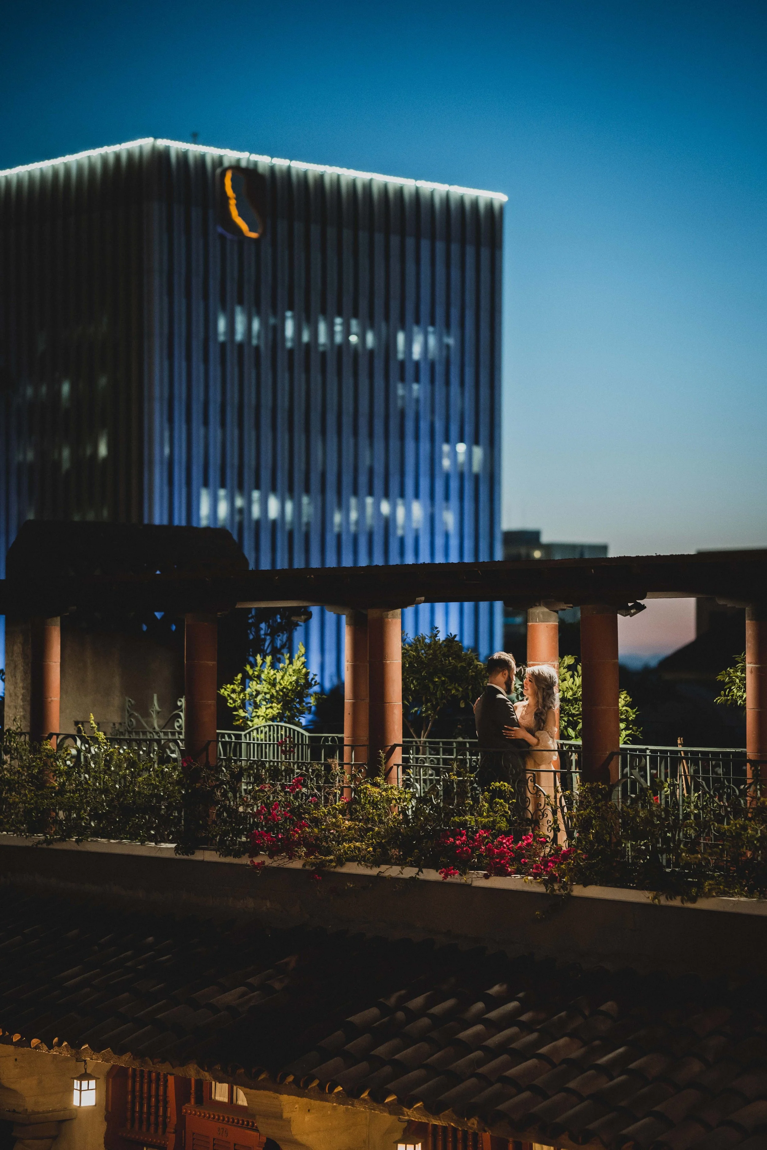 A couple stands together on a balcony at dusk, with a cityscape featuring a modern tall building illuminated with lights in the background, The Mission Inn in Riverside