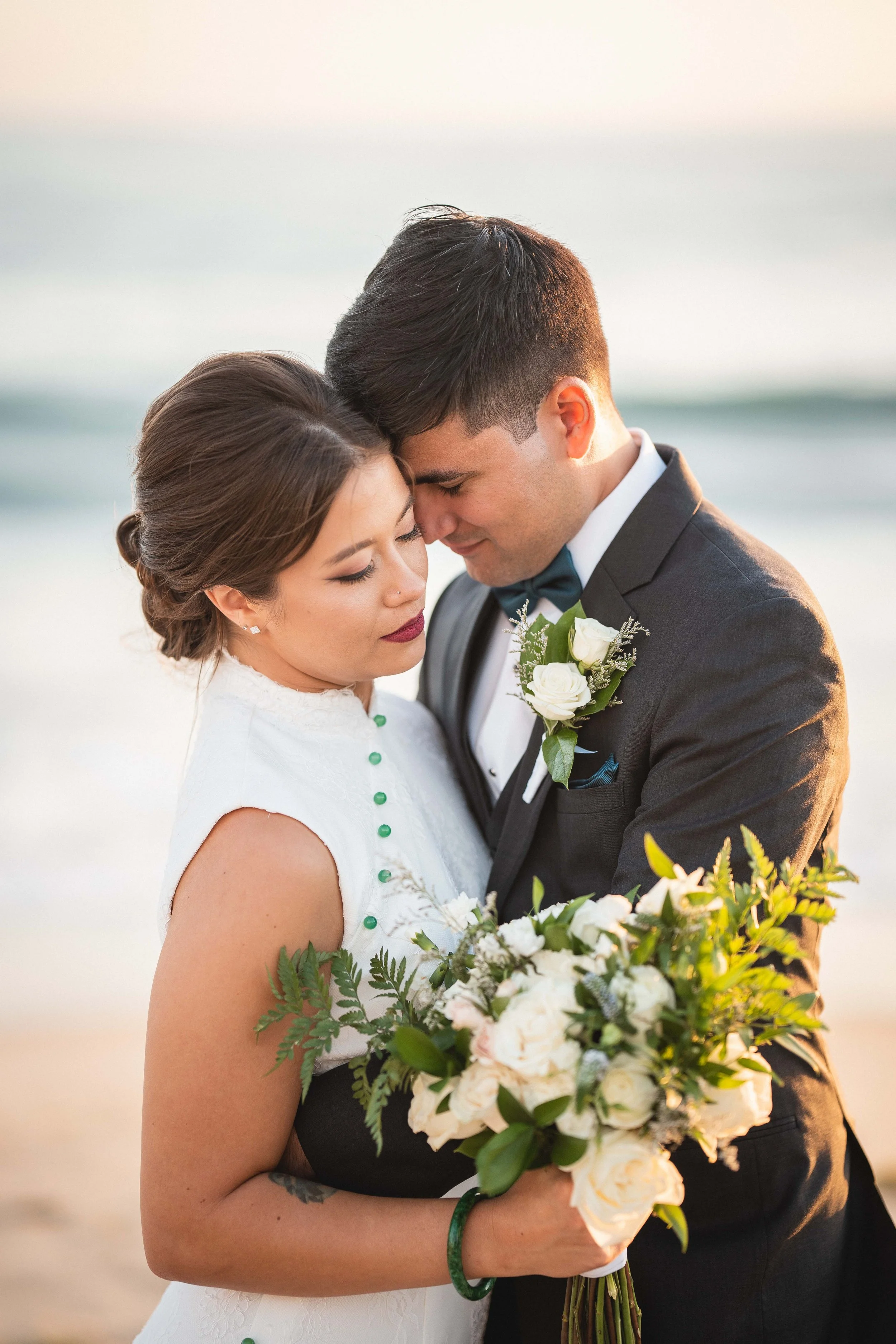 A bride and groom sharing a tender embrace on the beach at sunset, with the bride holding a bouquet of white flowers and greenery at Wedgewood San Clemente