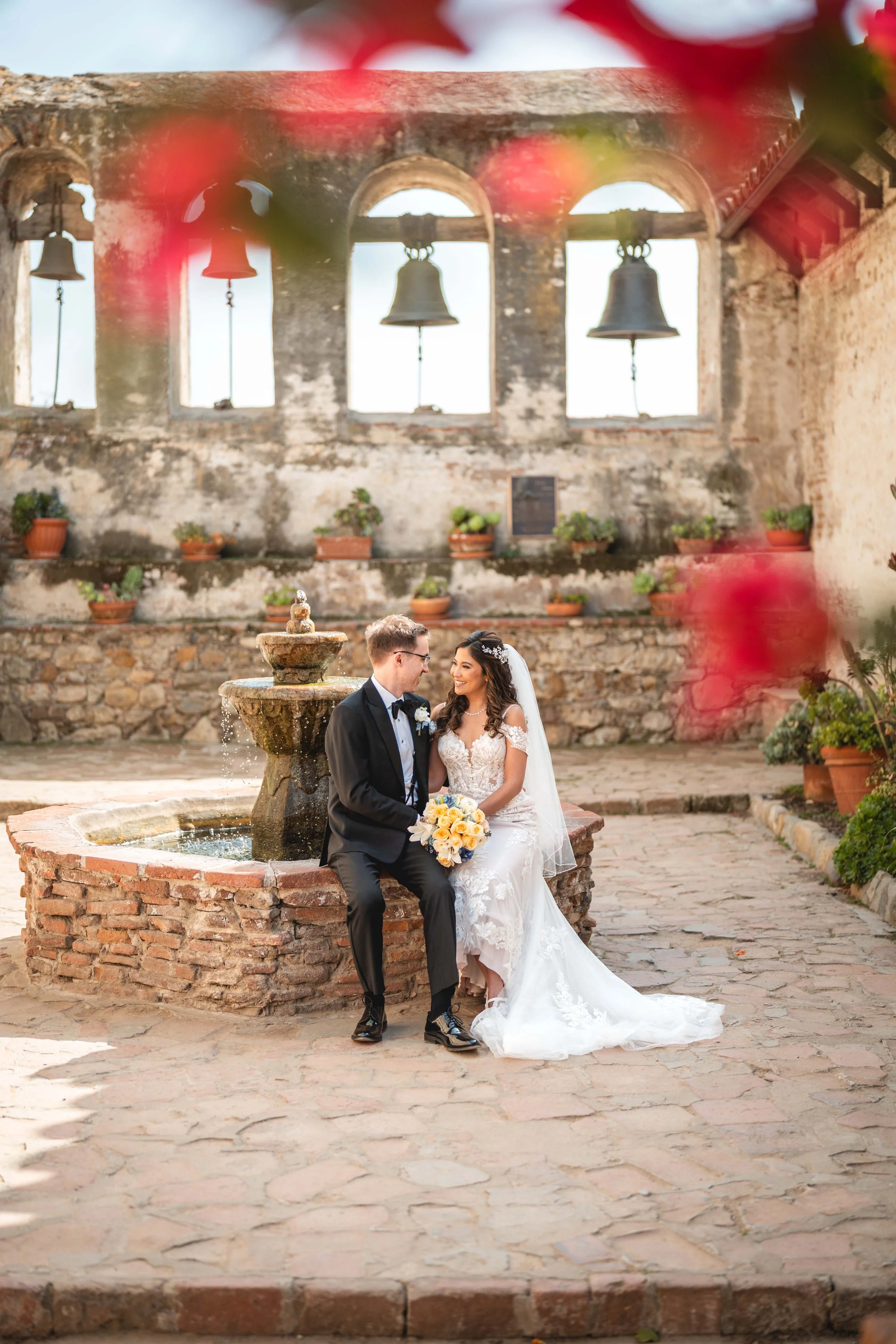 A newlywed couple sitting by a fountain in a rustic courtyard, with the bride holding a bouquet of yellow roses and both smiling at each other, Mission at San Juan Capistrano