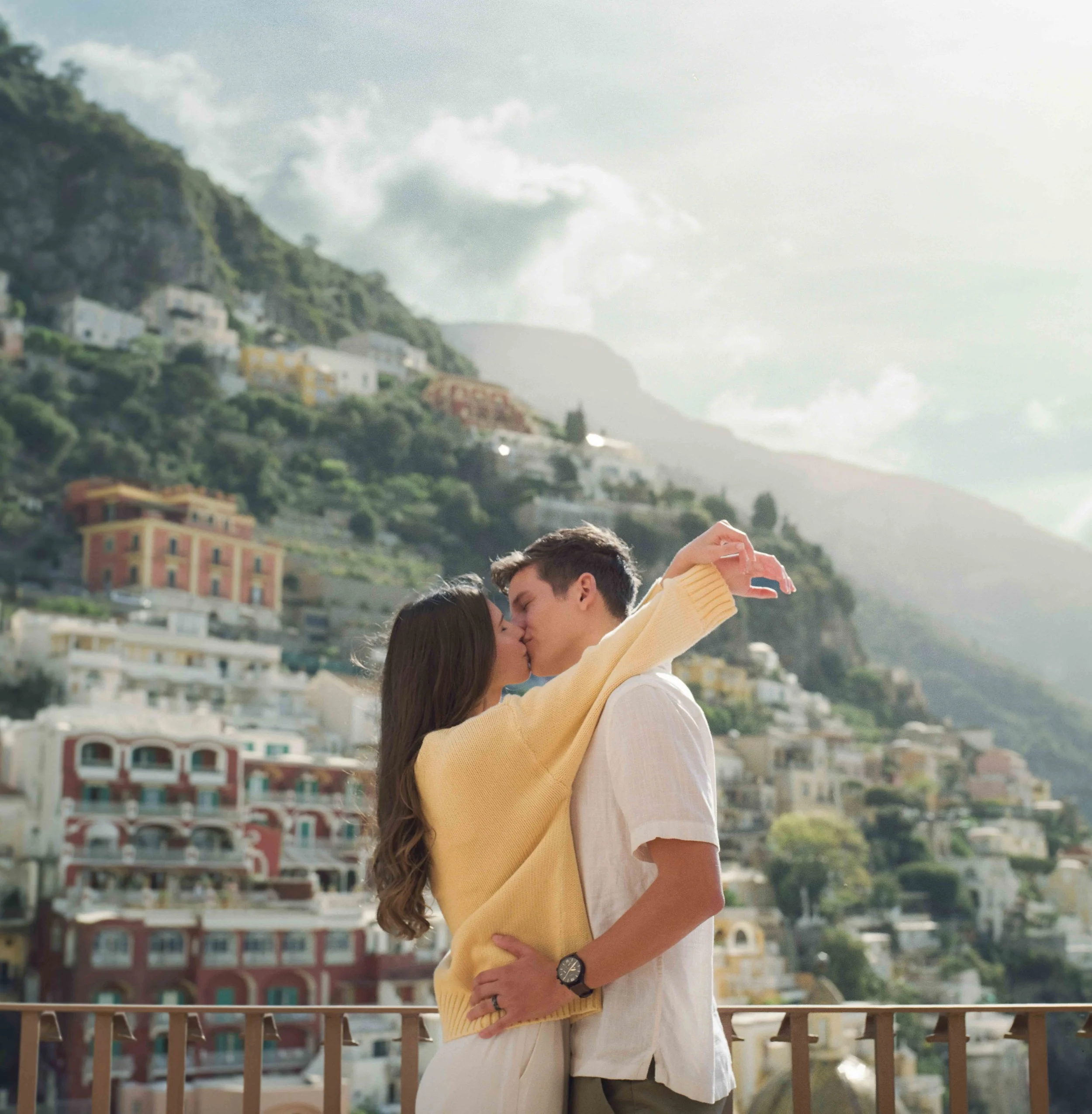 A couple sharing a kiss on a balcony with a hillside town in the background Positano Italy