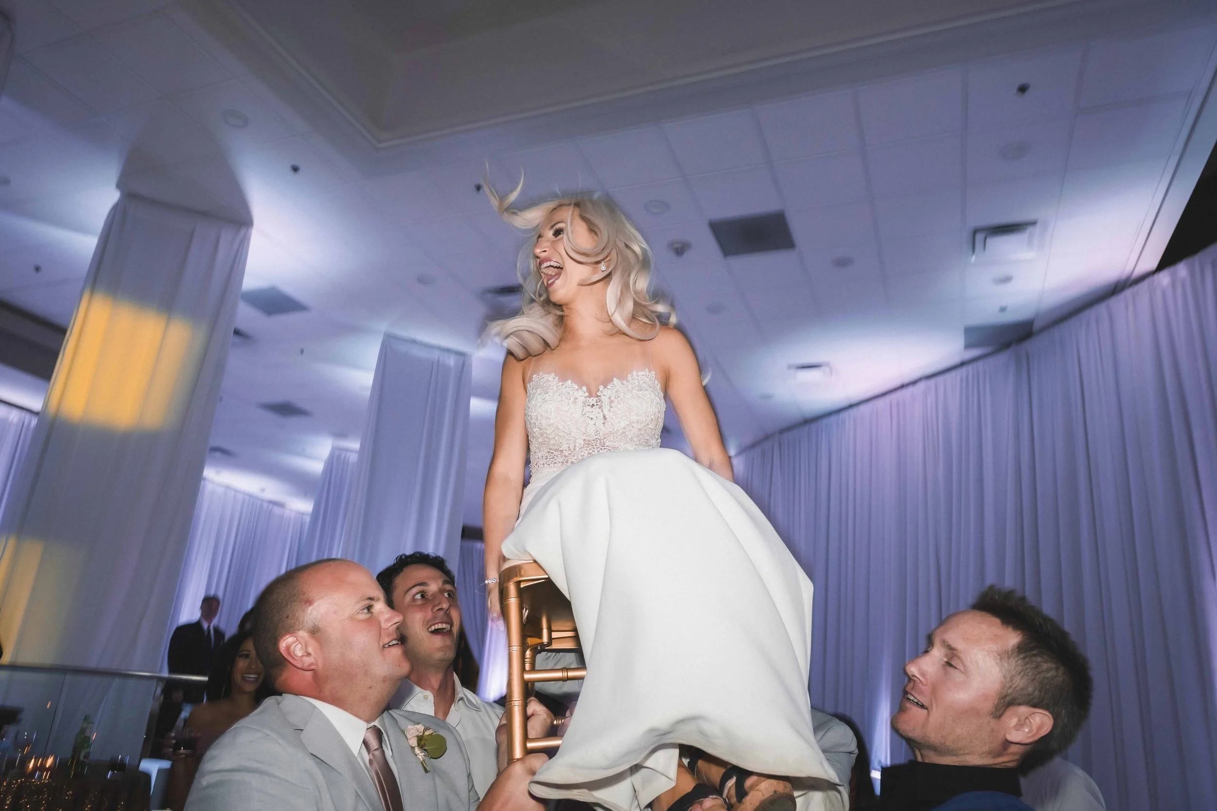 A bride in a white wedding dress is lifted on a chair by three men during a wedding celebration, all smiling and laughing inside a decorated venue with draped curtains and soft lighting, JW Marrott Palm Desert