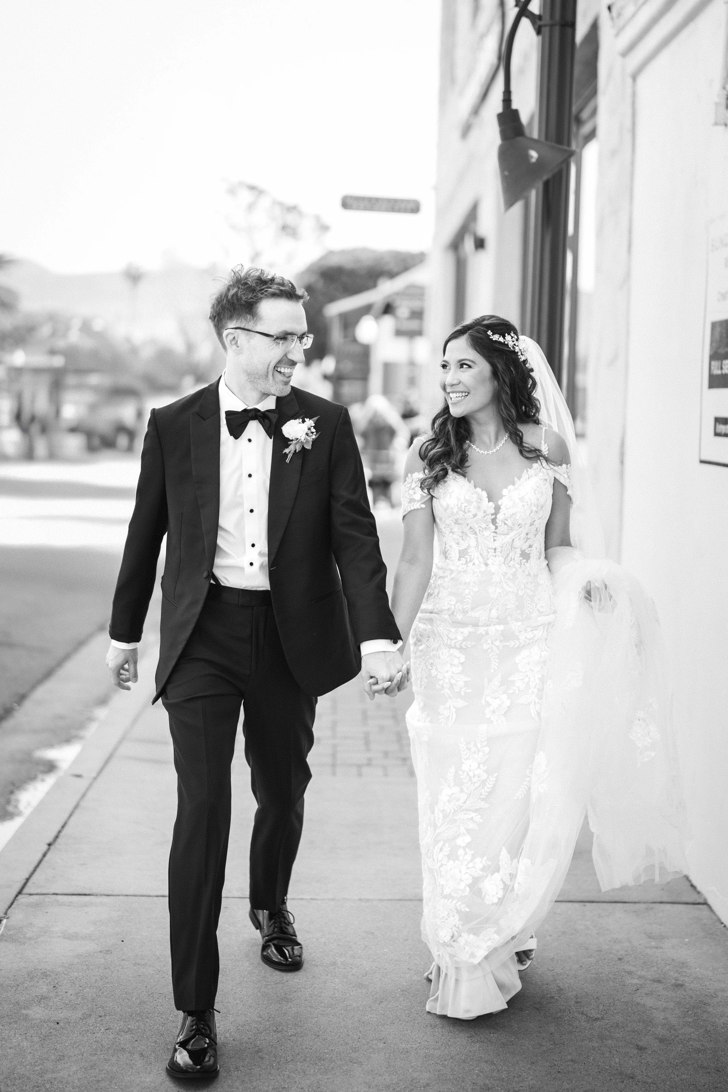 A black and white photo of a smiling bride and groom walking hand-in-hand on a sidewalk outside a building, dressed in wedding attire with the bride in a lace gown and the groom in a tuxedo franciscan gardens Mission San Juan Capistrano