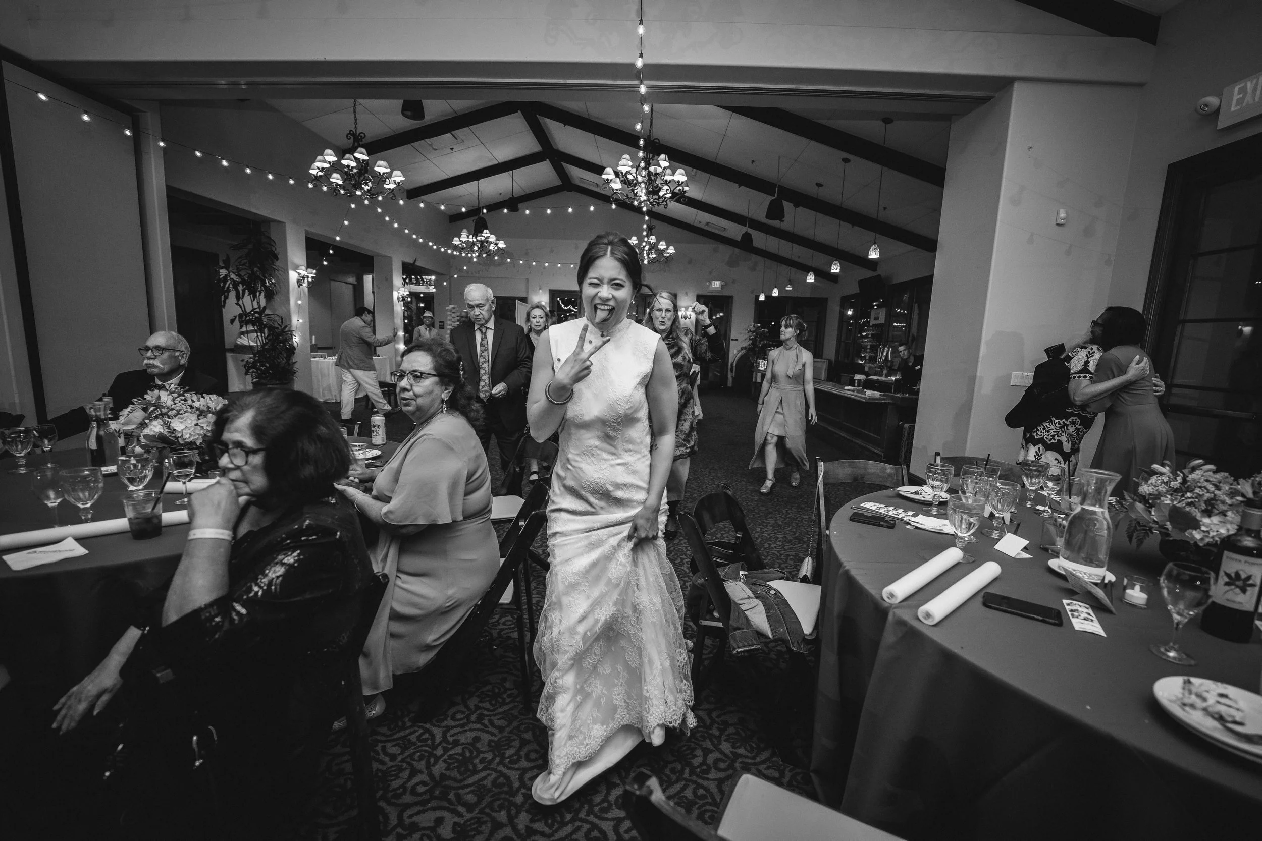 A group of people at a celebration or wedding reception, with one woman in a white dress smiling and making a peace sign, surrounded by others seated at tables and some standing and chatting, San Clemente Shores, Wedgewood