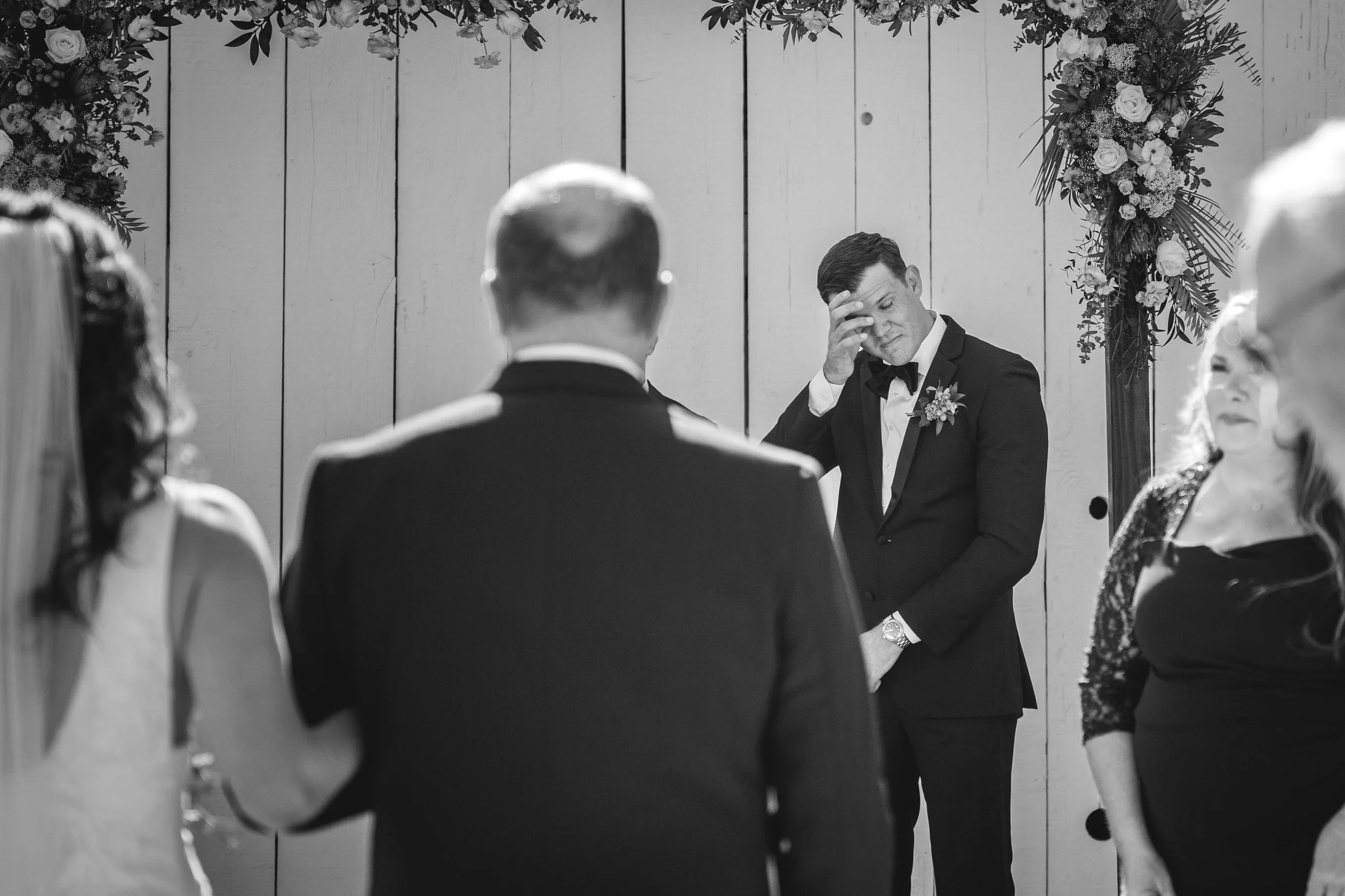 A black and white photo of a wedding ceremony with a groom and bride facing each other. The groom, in a suit, has his back to the camera. An officiant, in a tuxedo, is at the center, holding his forehead in a gesture of emotion, Aliso Viejo Ranch