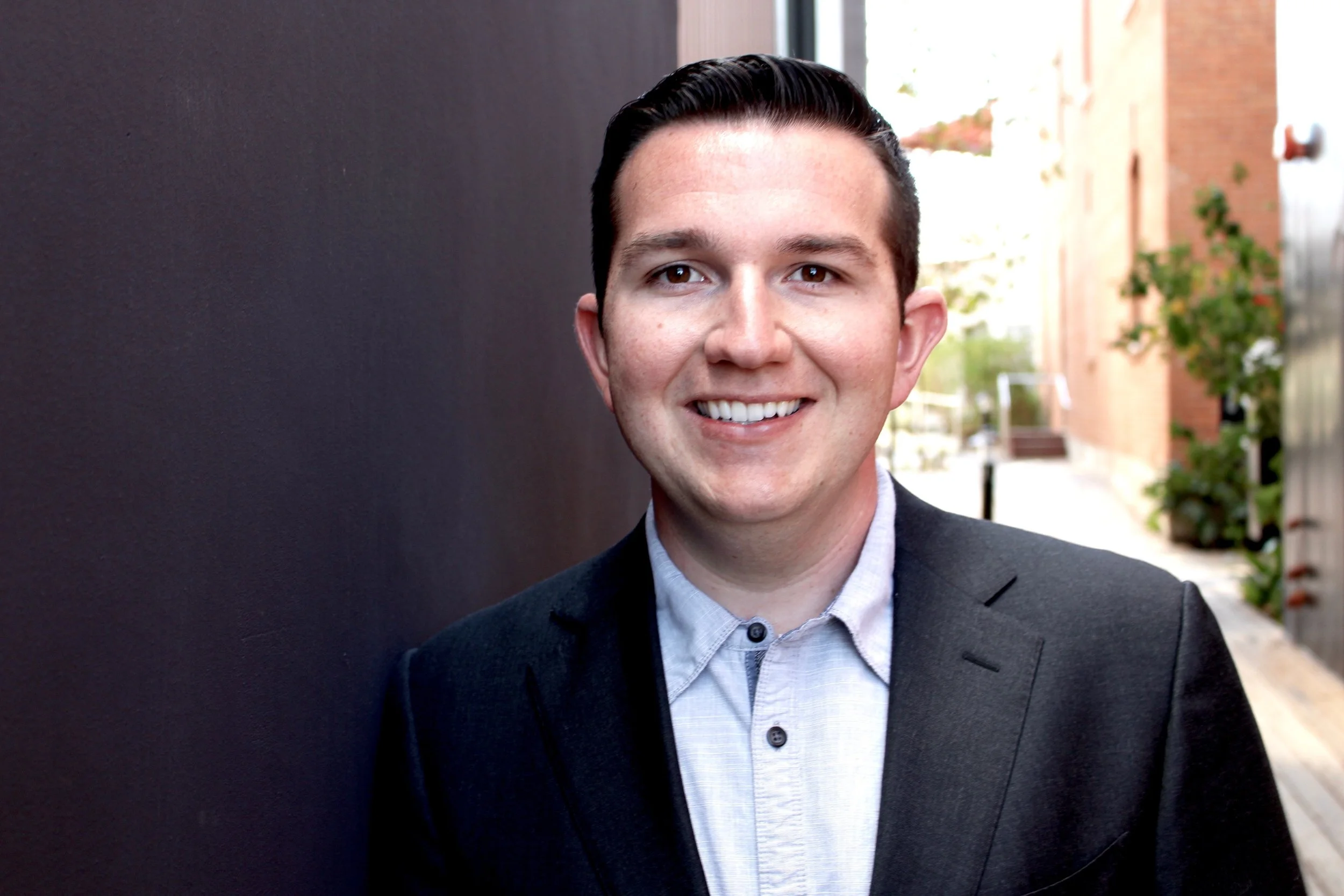 A young man with dark hair wearing a light gray button-up shirt and a black blazer, smiling outdoors against a dark wall with a building and trees in the background.