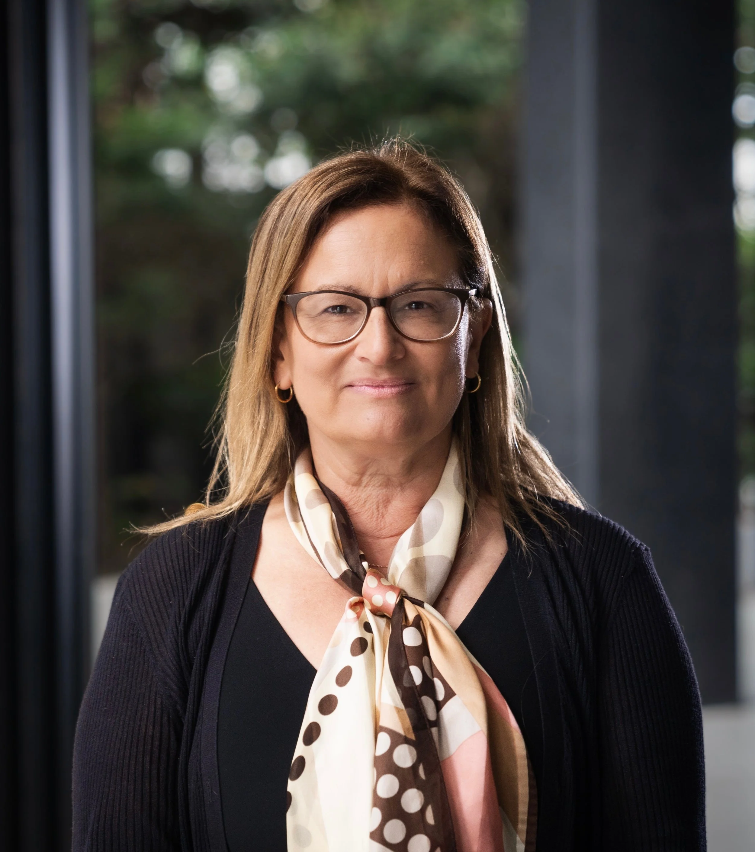 A middle-aged woman with glasses, wearing a black top and a polka dot scarf, standing indoors in front of a glass window with trees outside.