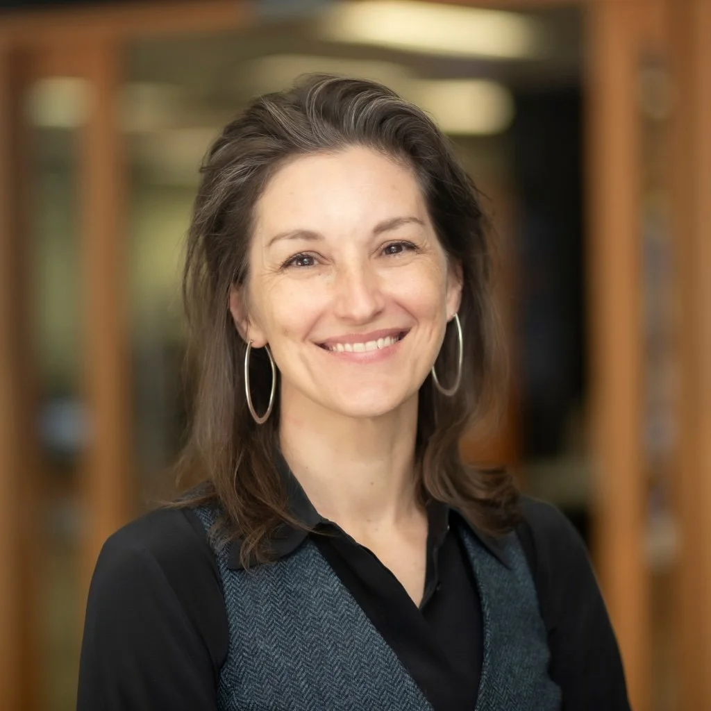 A woman with shoulder-length brown hair, wearing hoop earrings and a black top with a herringbone patterned vest, standing in front of a blurred bookshelf background, smiling at the camera.