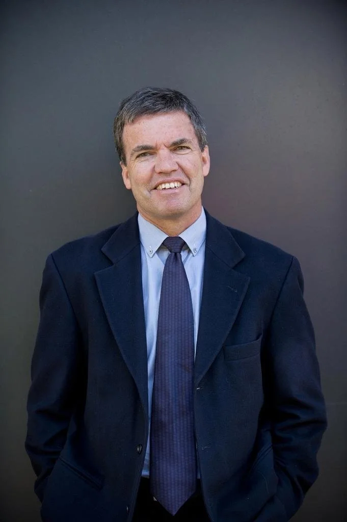 A man with short dark hair and a light complexion, wearing a dark suit, light blue shirt, and dark tie, standing against a plain dark gray background.