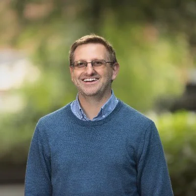 A man with glasses smiling outdoors, wearing a blue sweater and collared shirt, with blurred green foliage in the background.