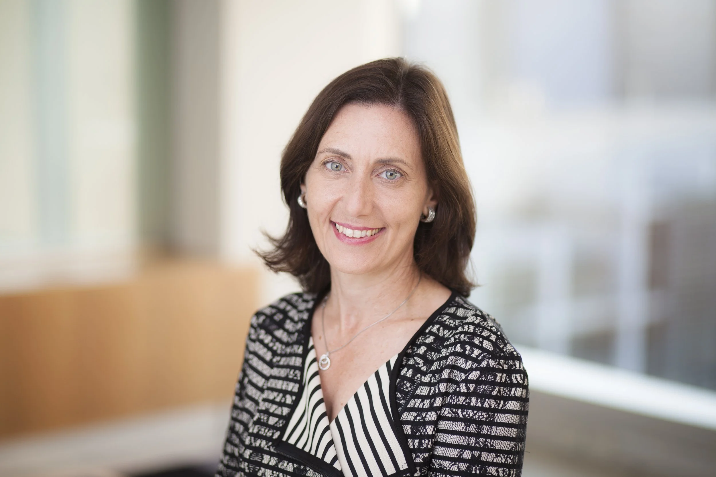 Professional woman with shoulder-length brown hair and blue eyes, wearing a black and white striped top with a black lace overlay, silver earrings, and a silver necklace, smiling in an indoor setting with blurred background.