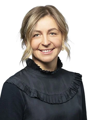 A young woman with blonde hair smiling, wearing a black high-neck blouse with ruffle detail.