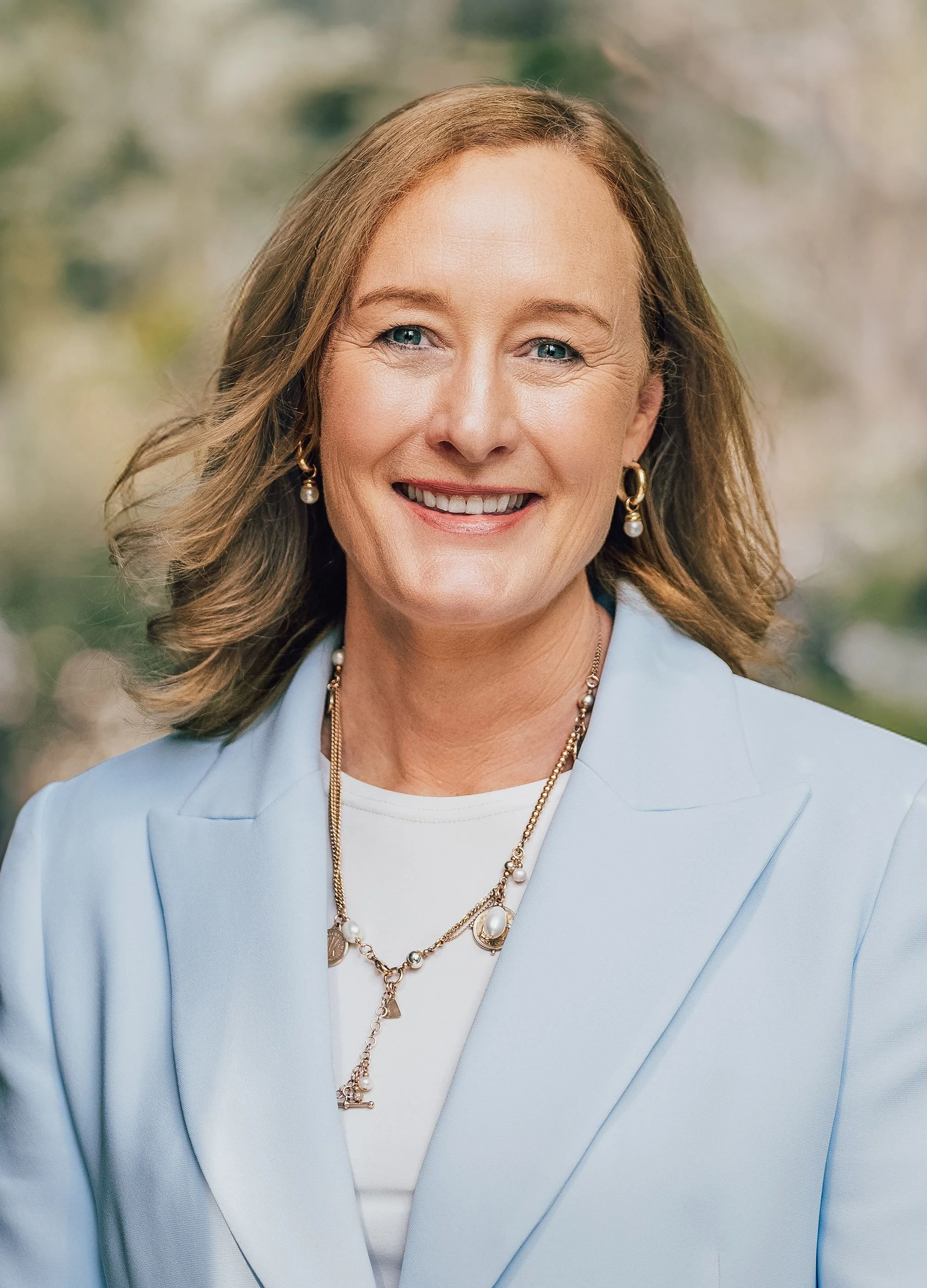 A portrait of a smiling middle-aged woman with shoulder-length light brown hair, wearing a light blue blazer and pearl jewelry, against a blurred outdoor background.
