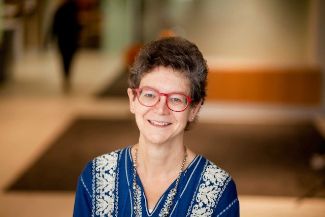 A middle-aged woman with short brown hair, red glasses, and a blue embroidered blouse smiling in an indoor setting.