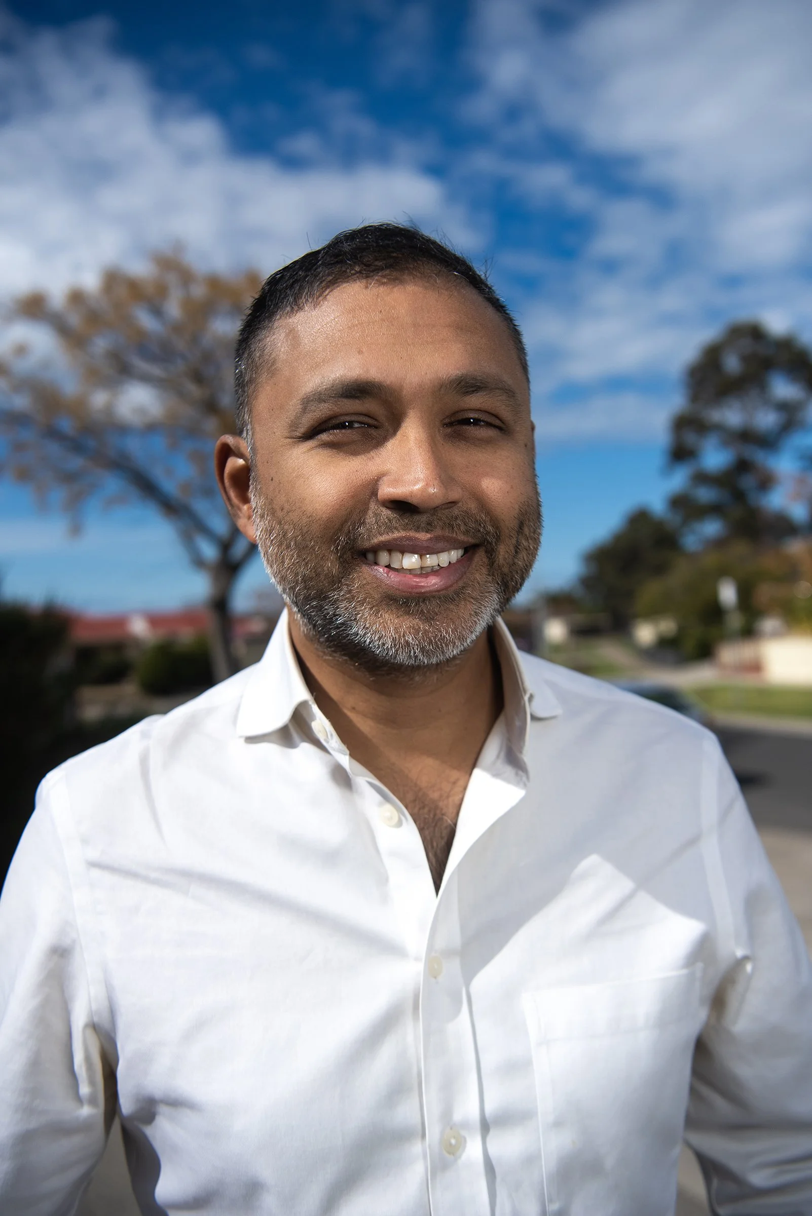 Man smiling outdoors with a blue sky and trees in the background.
