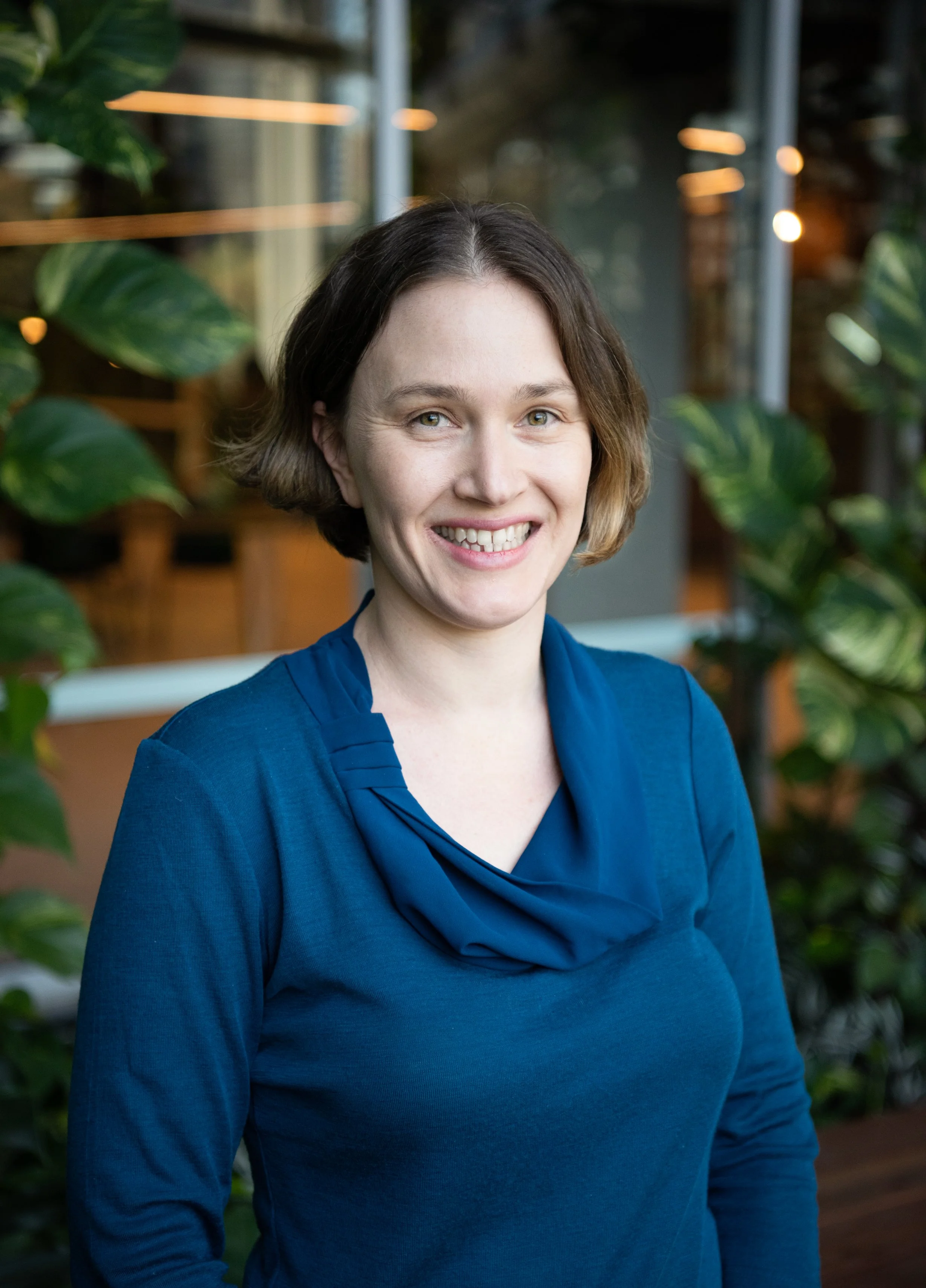 A woman with short brown hair, wearing a blue top with a cowl neck, smiling, standing in front of green plants and a glass window in an indoor setting.