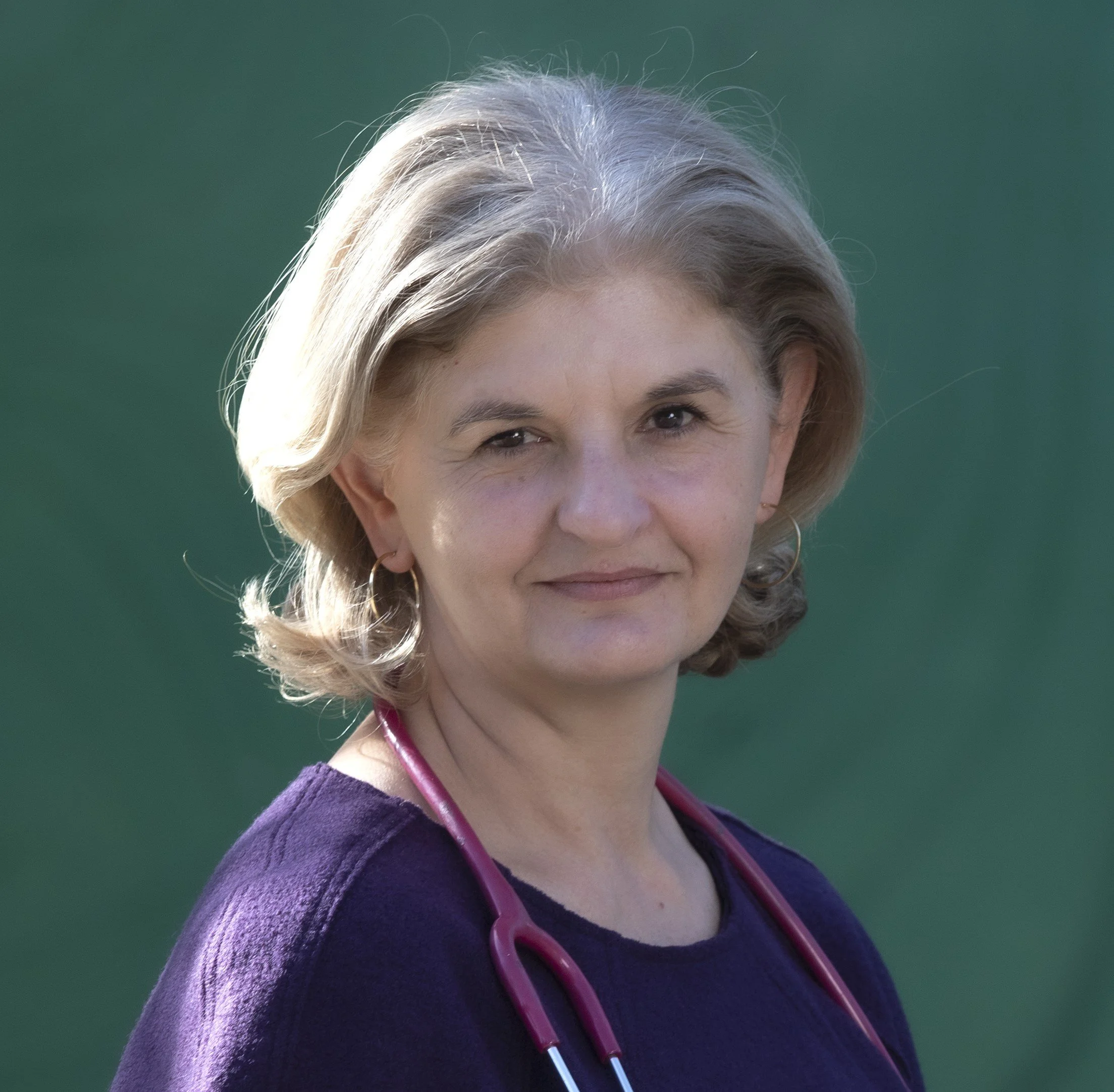 Portrait of a middle-aged woman with blonde hair, wearing a purple top and a pink stethoscope around her neck, standing outdoors against a green background.